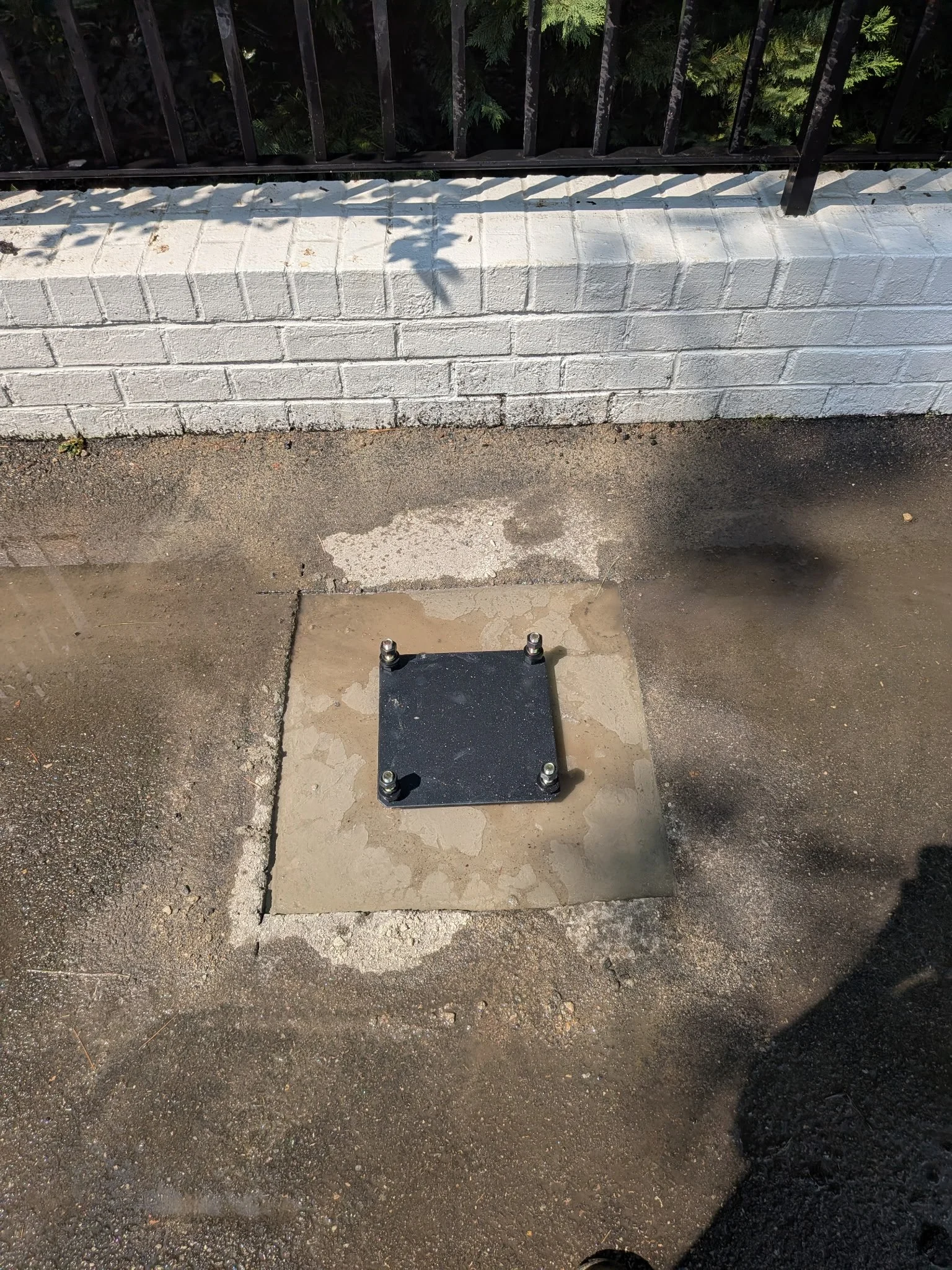 Black metal square plate with four bolts on a concrete sidewalk in front of a white brick wall and a black metal fence with green foliage behind.