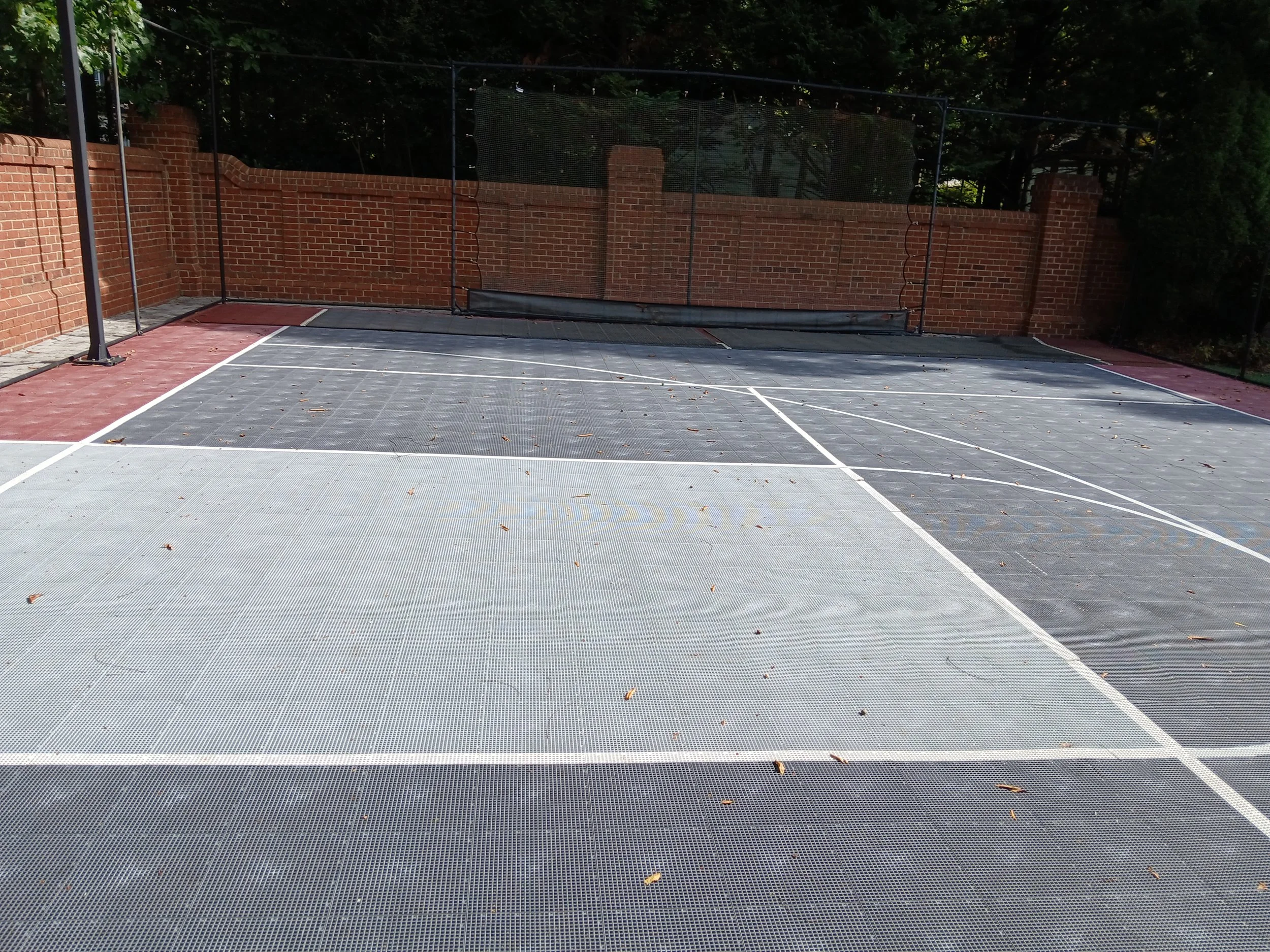 Empty outdoor tennis court with a brick wall, green trees, and a black net in the background.