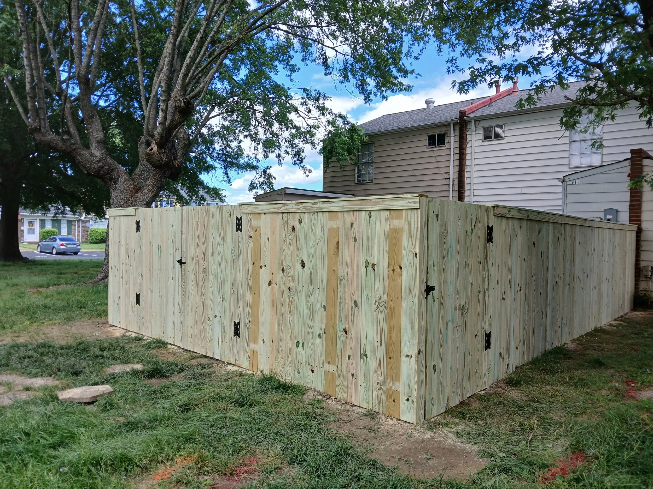 A newly built wooden fence surrounds a backyard, with a large tree and neighboring houses in the background.