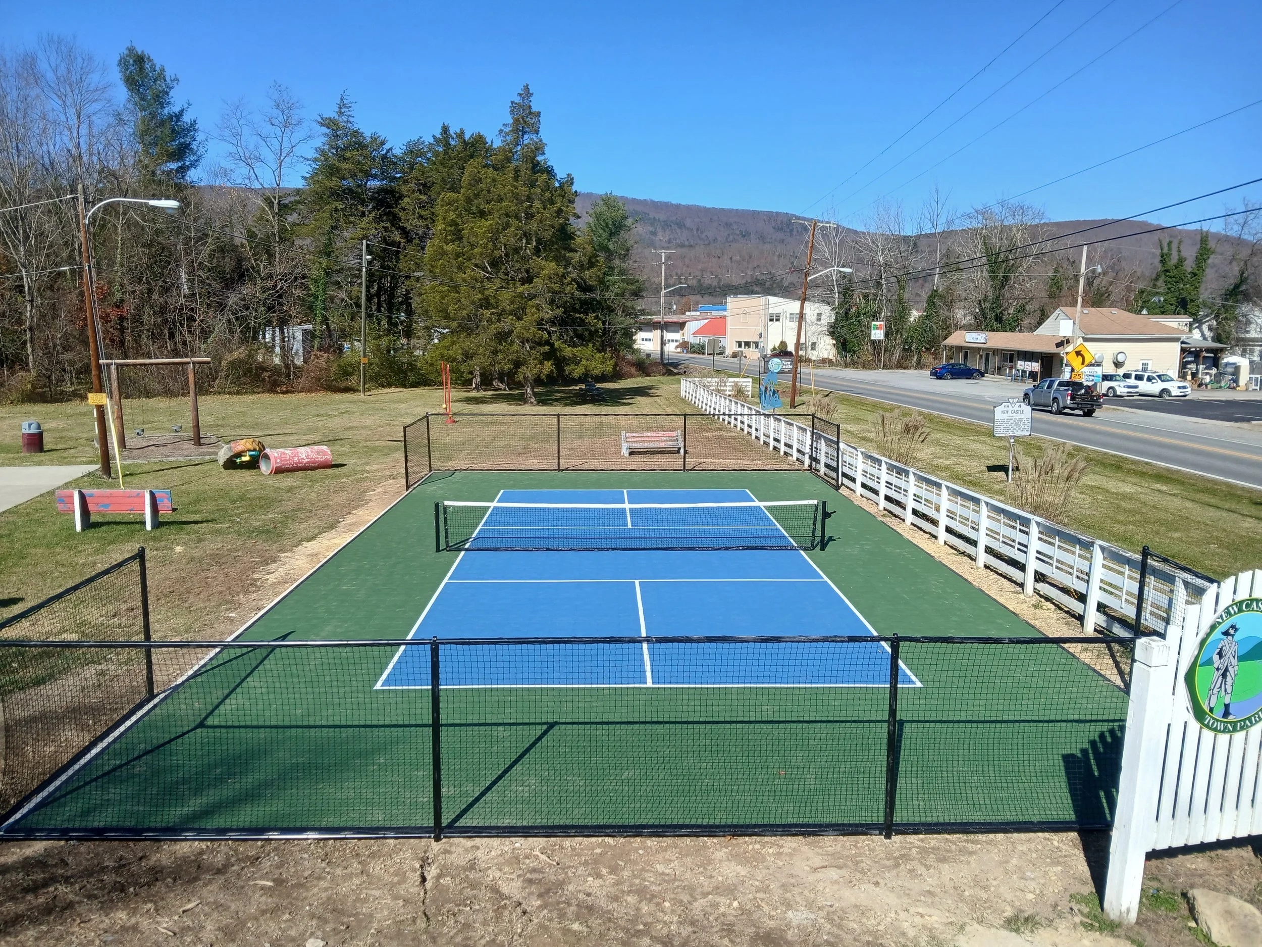 Small outdoor tennis court with a blue playing surface, surrounded by a black fence, with a white picket fence along one side. In the background, there is a grassy area with trees, playground equipment, and nearby buildings along a road with cars and