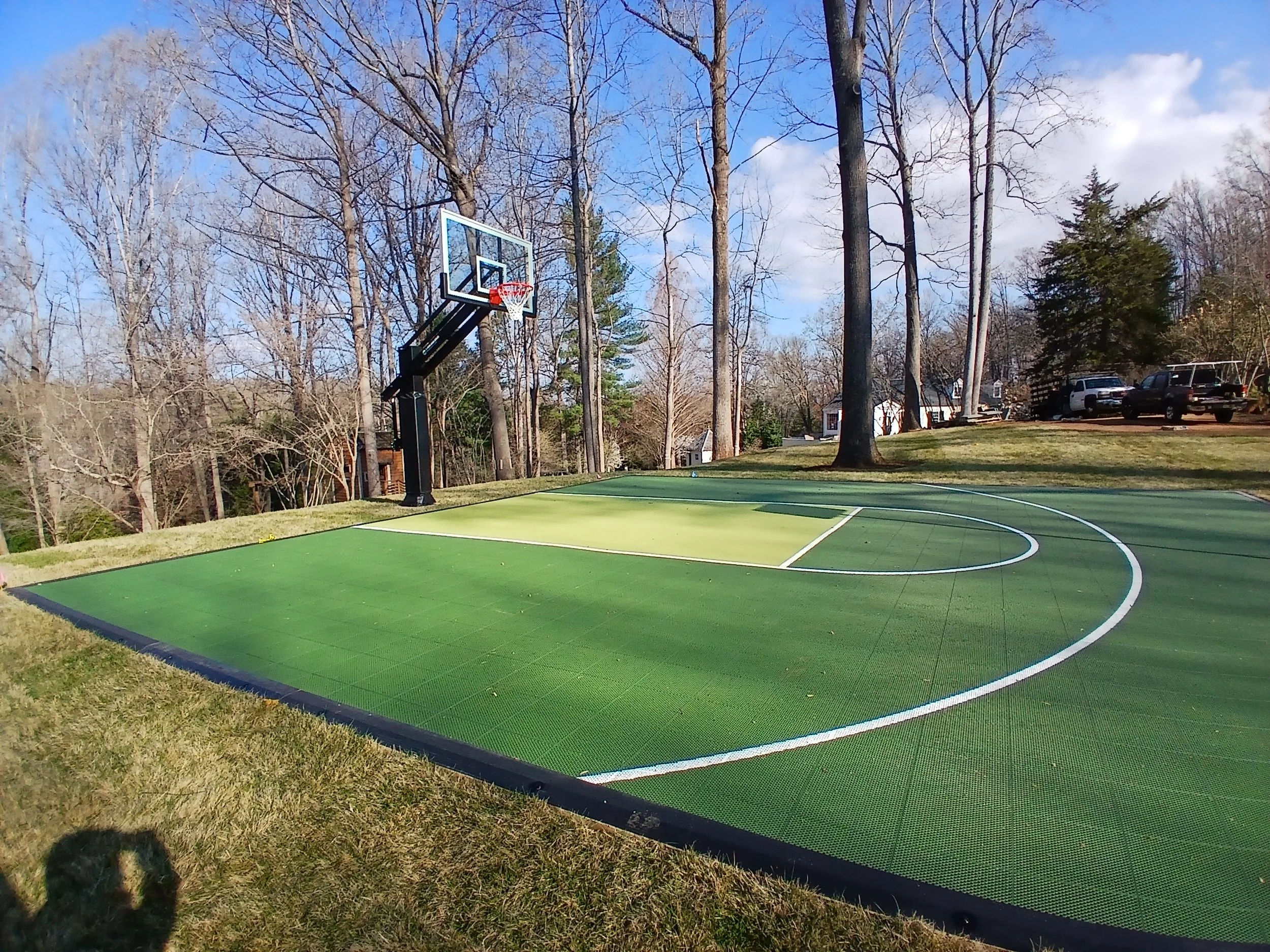 An outdoor basketball court with a green playing surface, a basketball hoop, and a wooded background with leafless trees and parked cars.