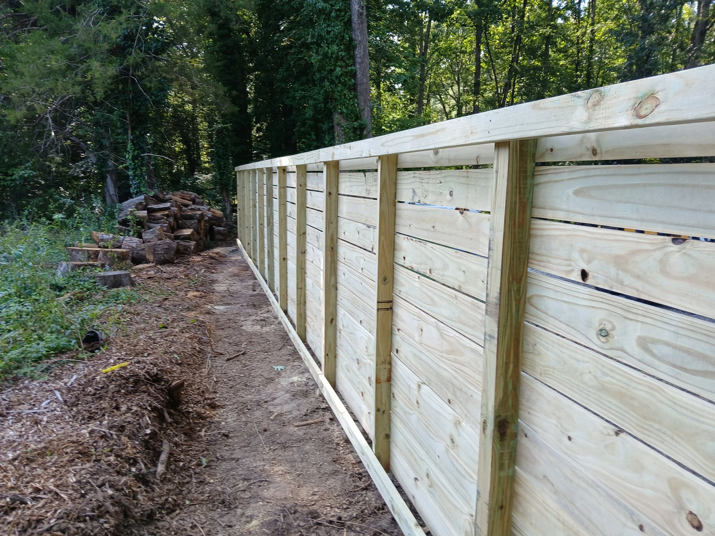 Wooden fence being installed along a forest trail, with a pile of logs nearby.