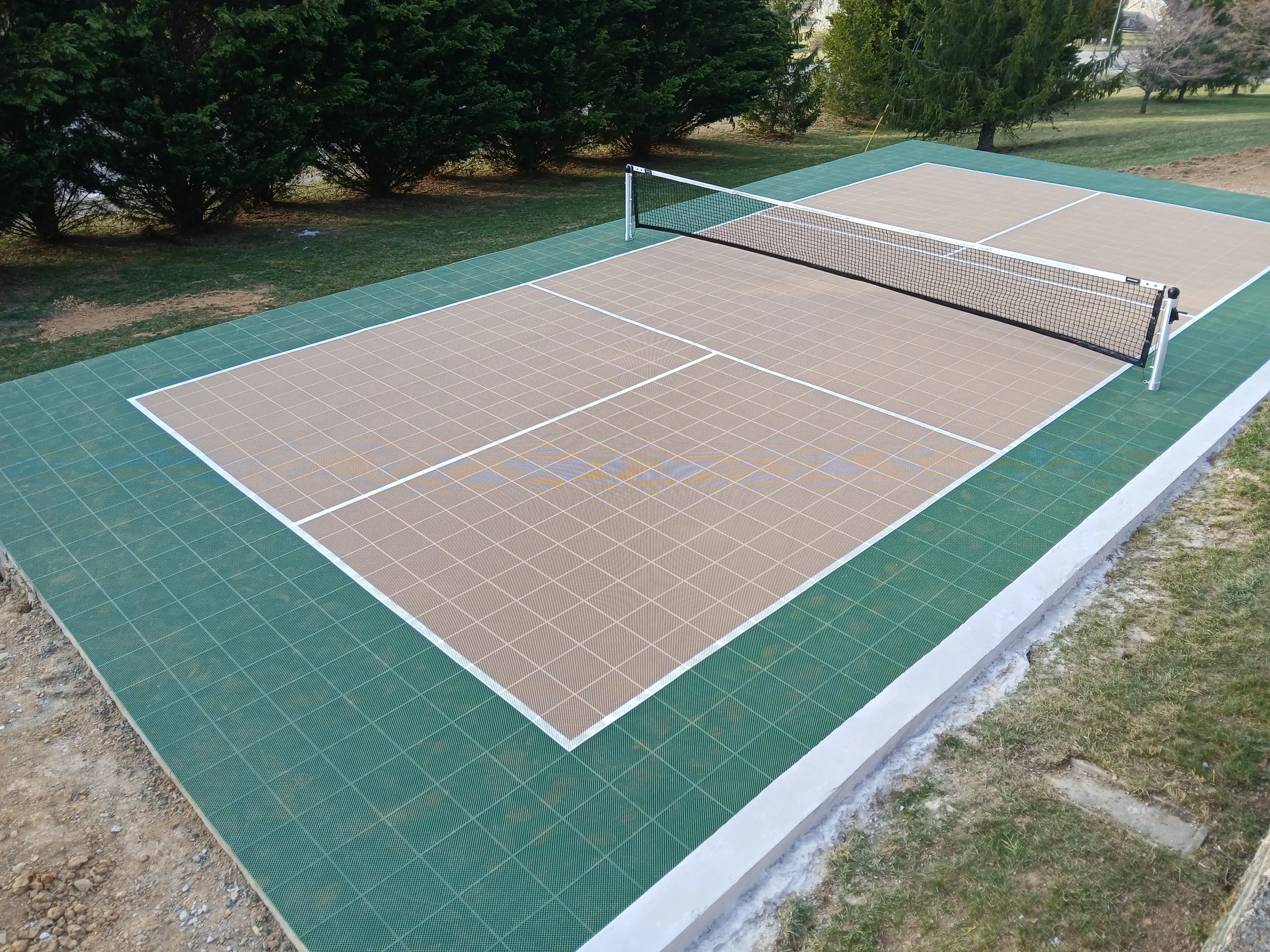 Outdoor pickleball court with a tan surface, green border, and black net, surrounded by grass and trees.