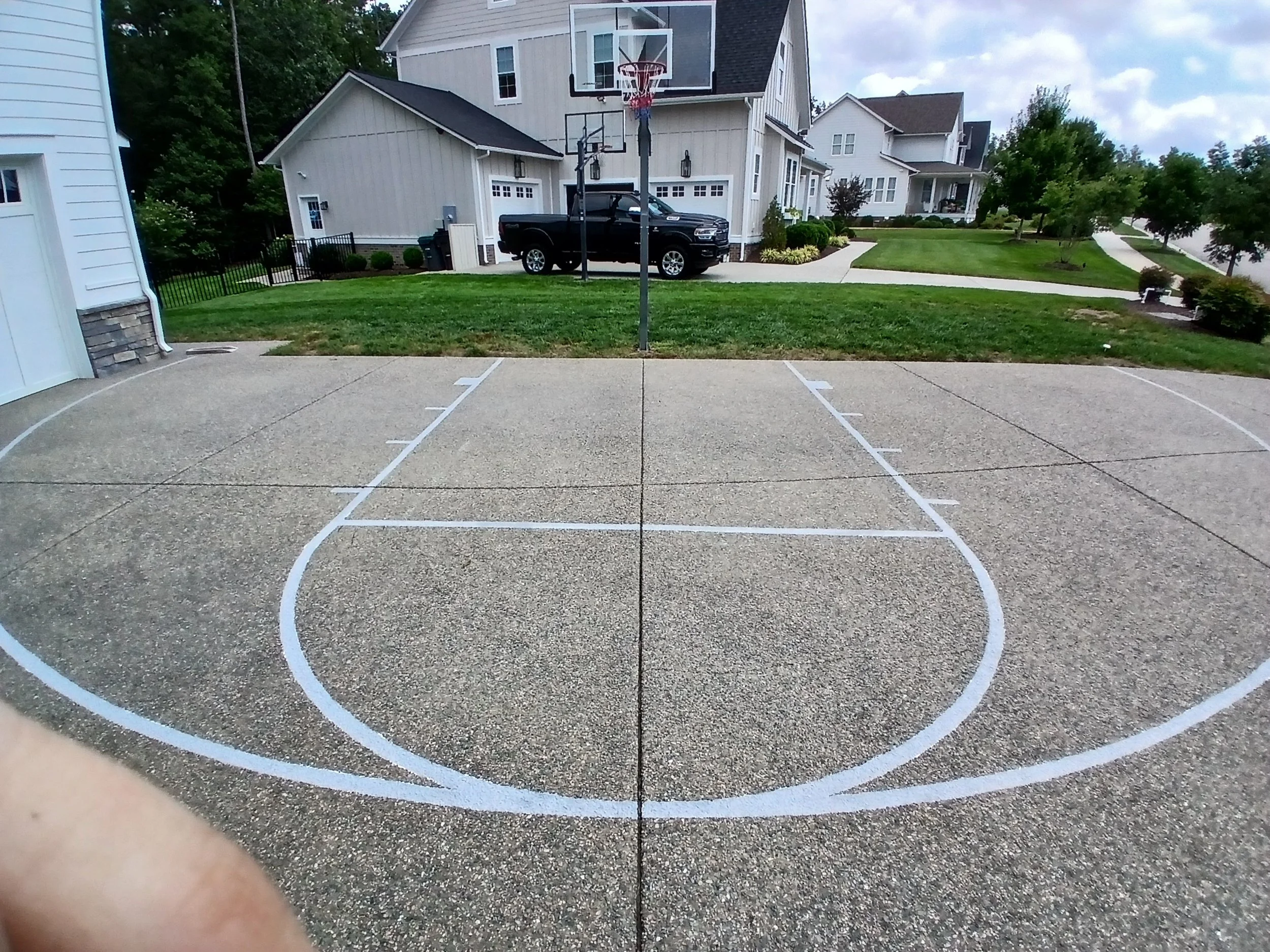 Empty outdoor basketball court with white painted lines on concrete, surrounded by residential houses and green lawns.