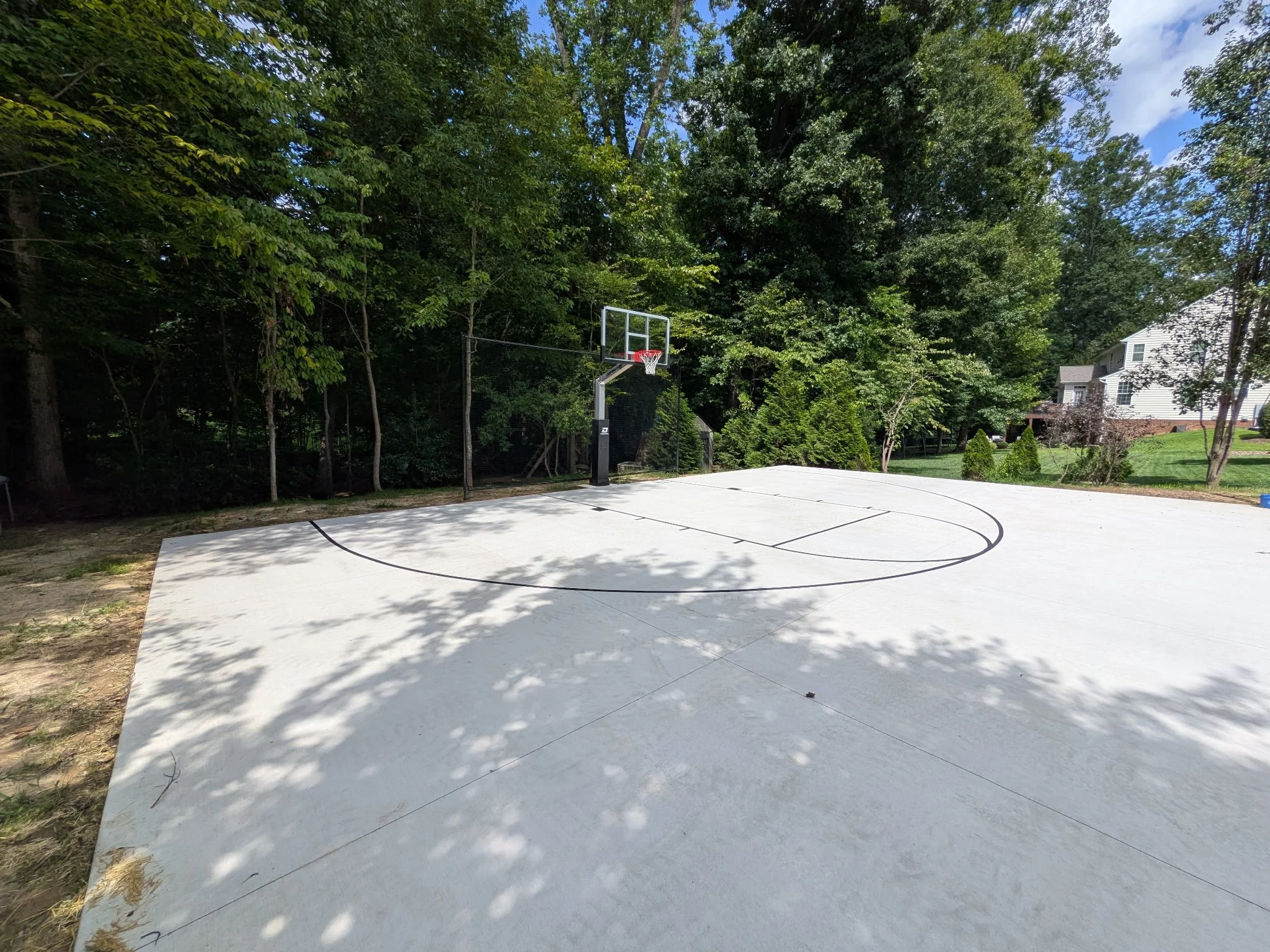 An outdoor basketball court with a basketball hoop and backboard, surrounded by trees and residential houses, with shadows cast on the court.