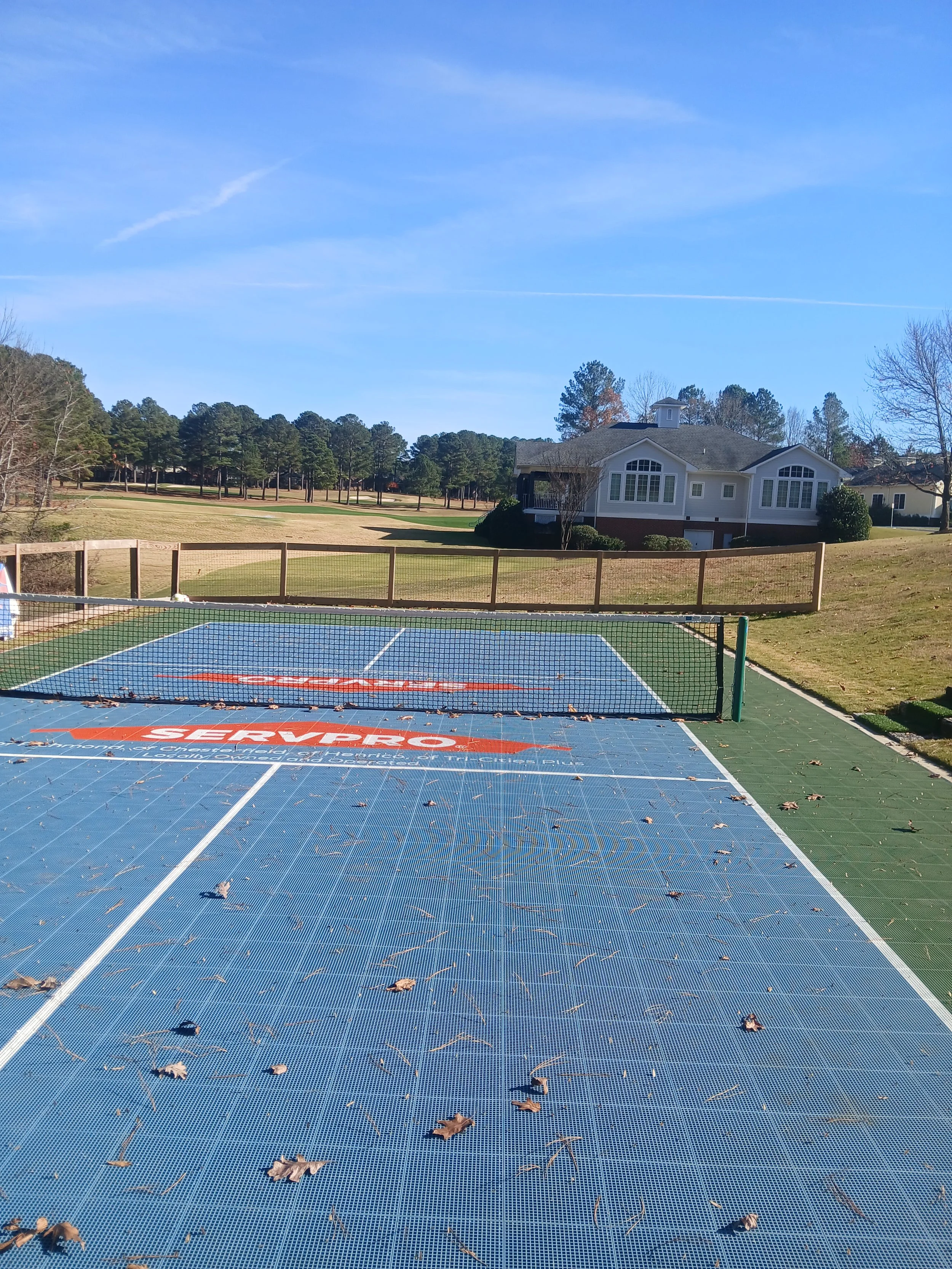 Outdoor tennis or pickleball court with a fallen leaves scattered on the ground, surrounded by a safety fence, with houses and trees in the background under a blue sky.