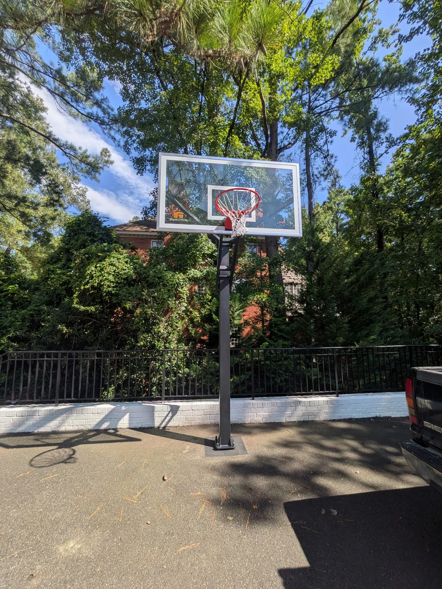 An outdoor basketball hoop standing on the asphalt court with a green fence, trees, a brick house, and blue sky in the background.