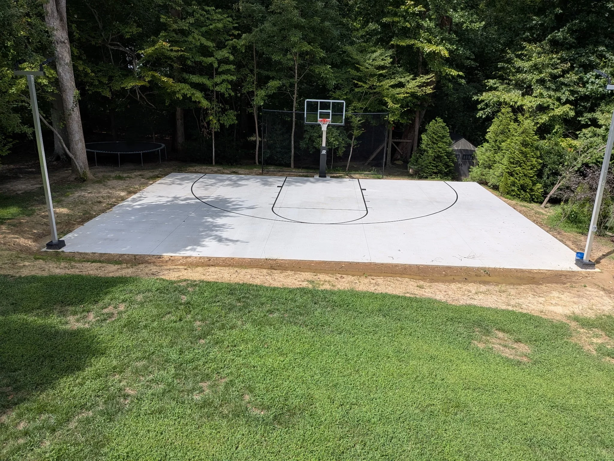 An outdoor basketball court with a concrete surface and a basketball hoop, surrounded by trees and greenery.