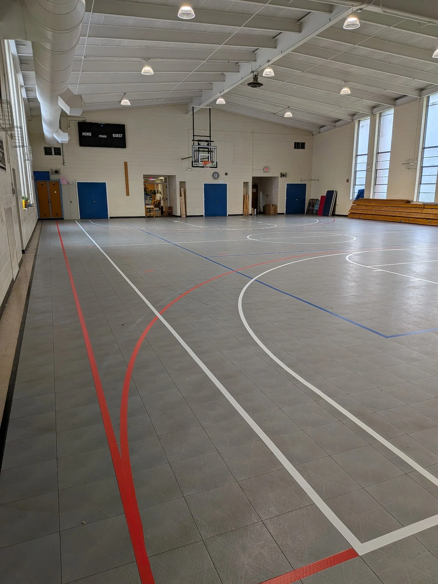 An indoor gymnasium with a basketball hoop, benches, lockers, and high windows, with a scoreboard on the wall.