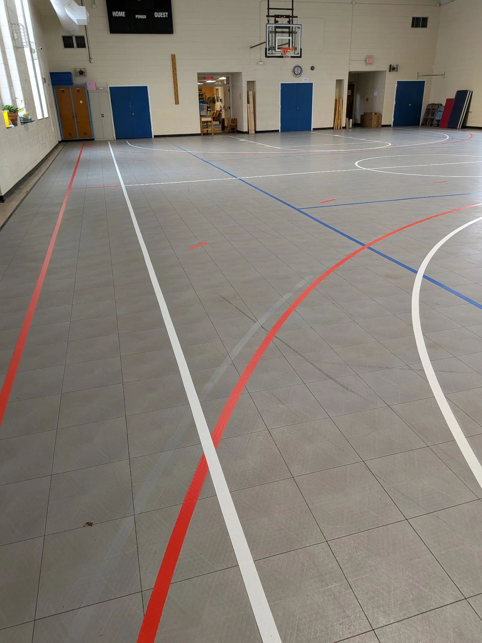 Empty indoor gymnasium with basketball hoop, scoreboard, and blue doors along the wall.