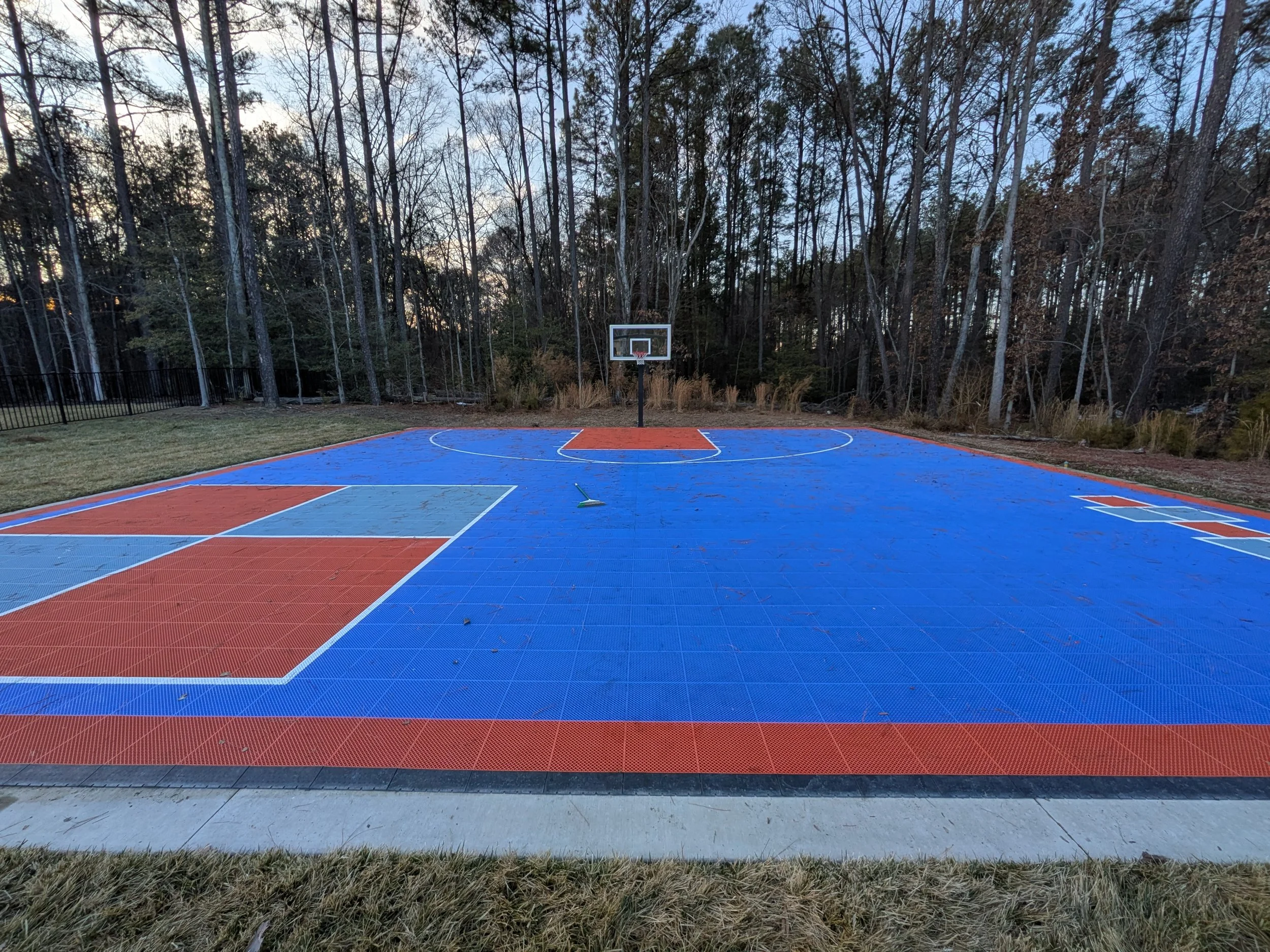 An outdoor basketball court with blue and orange surface, surrounded by a grassy area and trees, with a basketball hoop at the far end.