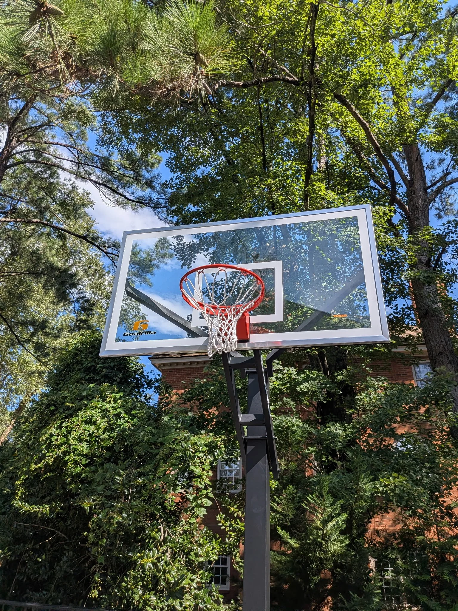 A basketball hoop with a backboard, orange rim, and white net is set outdoors among green trees and a brick building in the background.