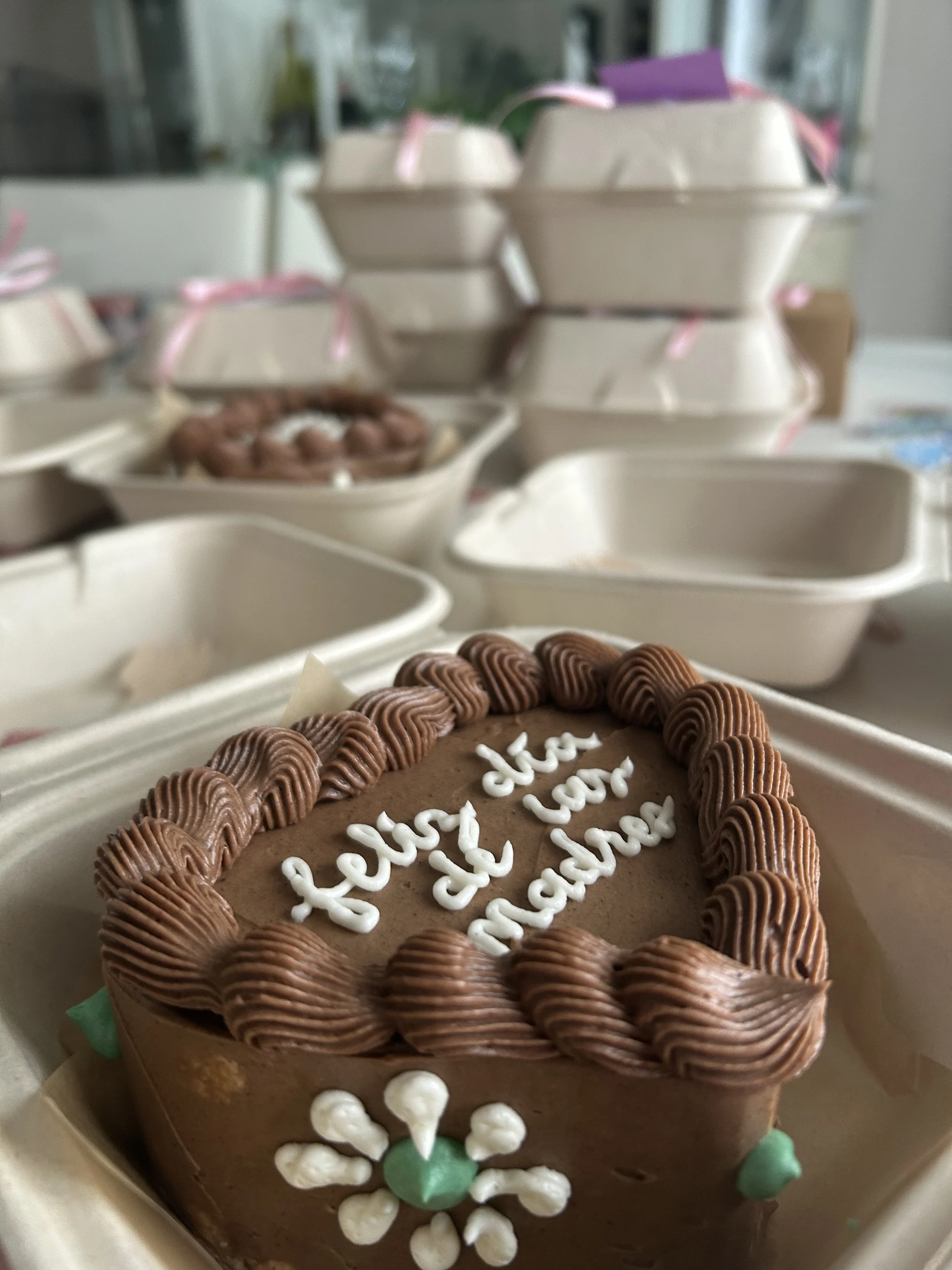 A decorated chocolate cake with white writing and brown piping, surrounded by empty white takeout containers and packages, at an event or celebration.