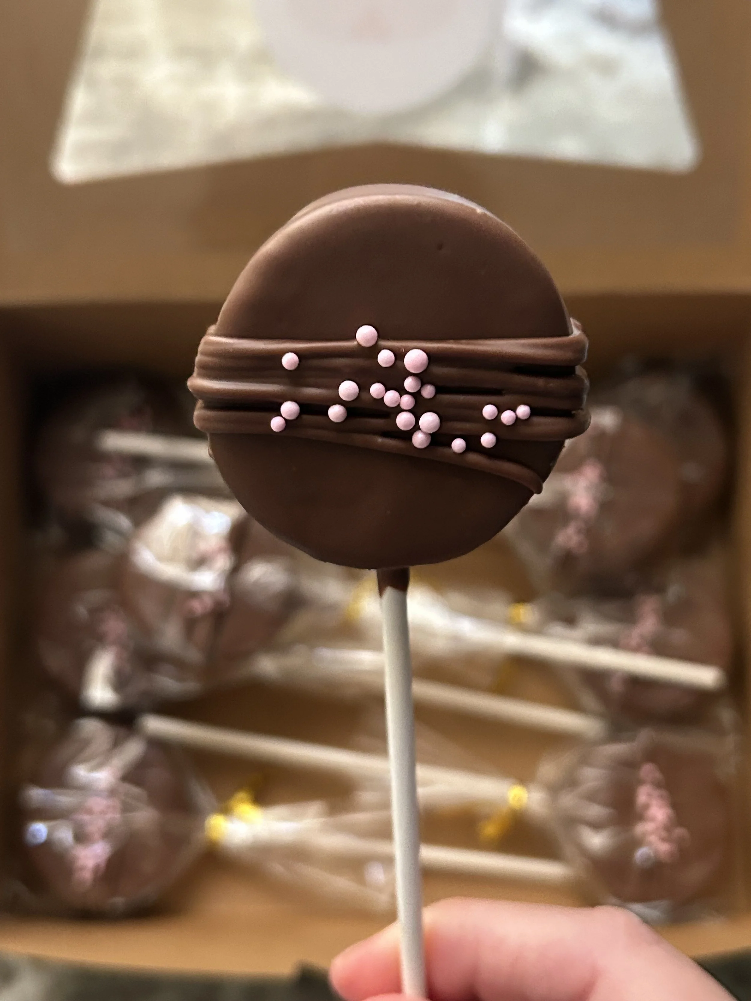 A close-up of a chocolate lollipop with pink sprinkles, held on a stick, with a box of similar treats in the background.