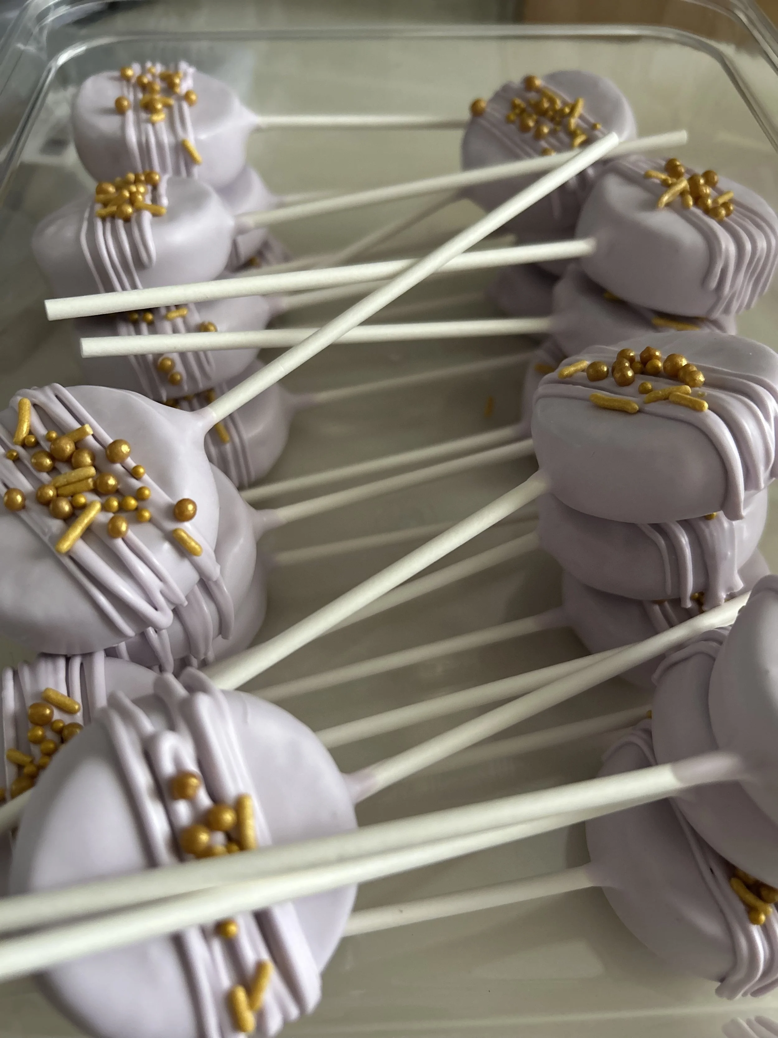 Lemon meringue pops with white coating, topped with gold sprinkles, on white sticks arranged in a clear container.