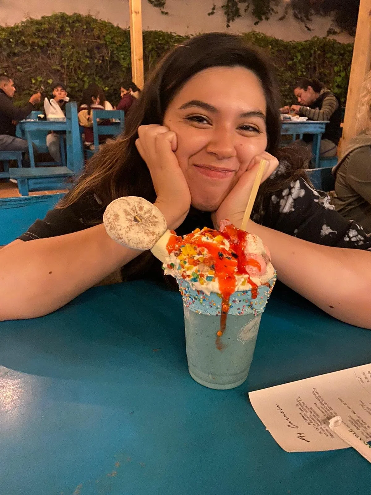 A young woman with dark hair smiling with her chin resting on her hands, sitting at a blue table in a restaurant or cafe. In front of her is a large milkshake topped with whipped cream, colorful sprinkles, and caramel syrup, with a cookie and a piece of marshmallow on the rim. There are other people sitting at tables in the background.