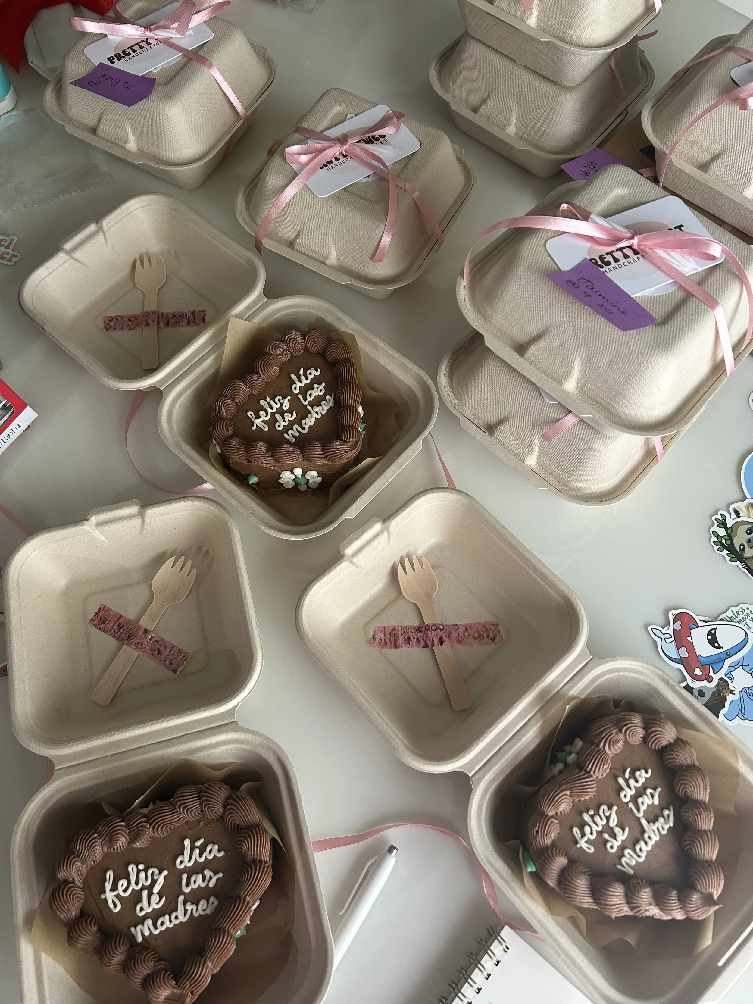 Heart-shaped chocolate cakes with white writing that says 'feliz día de las madre', surrounded by beige eco-friendly food containers with pink ribbons, small wooden forks, and decorative stickers for Mother's Day celebration.