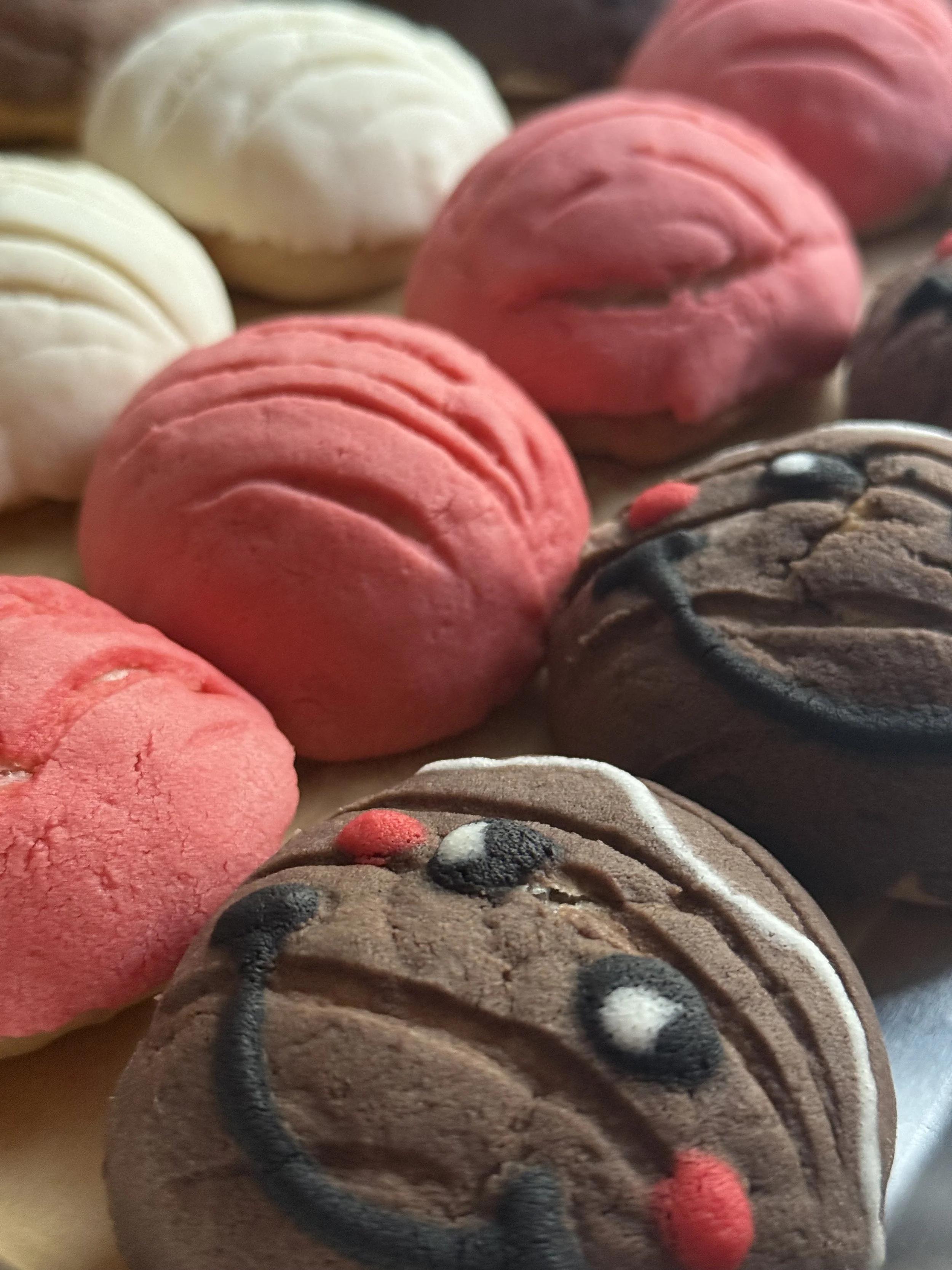 Close-up of cookies decorated to resemble smiling faces, with pink, white, and chocolate colors, some with red and white dots and black curved lines for the mouth and eyes.