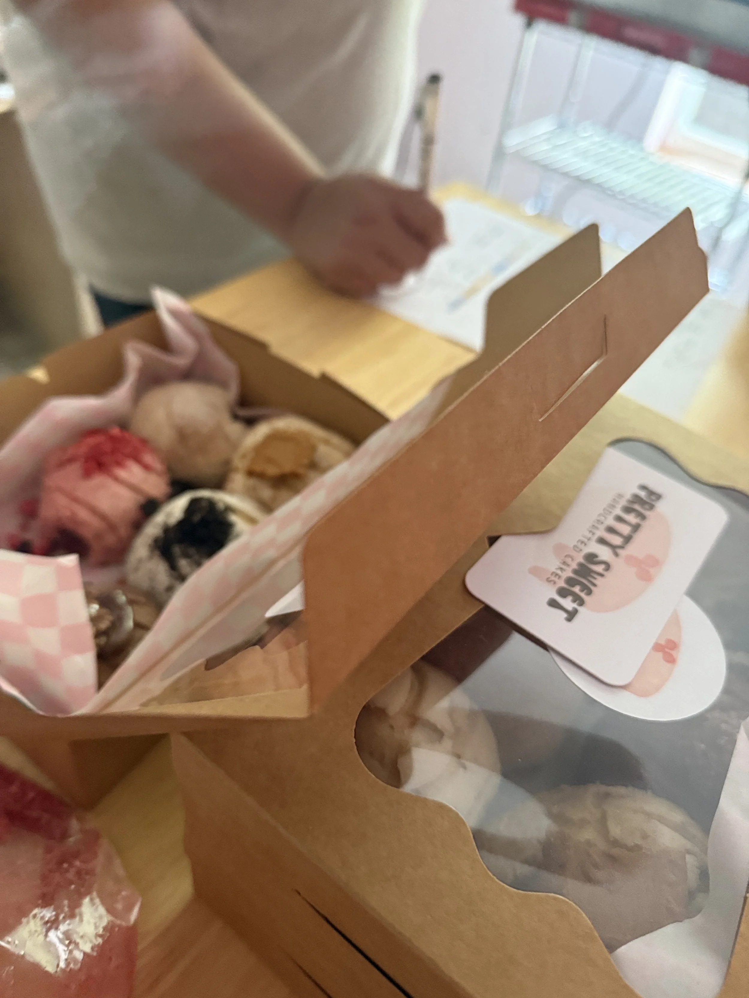 A hand holding a box of assorted cupcakes with pink and white checkered lining, on a table near a person writing on a notepad, with a sticker reading 'Party Sweet' visible.