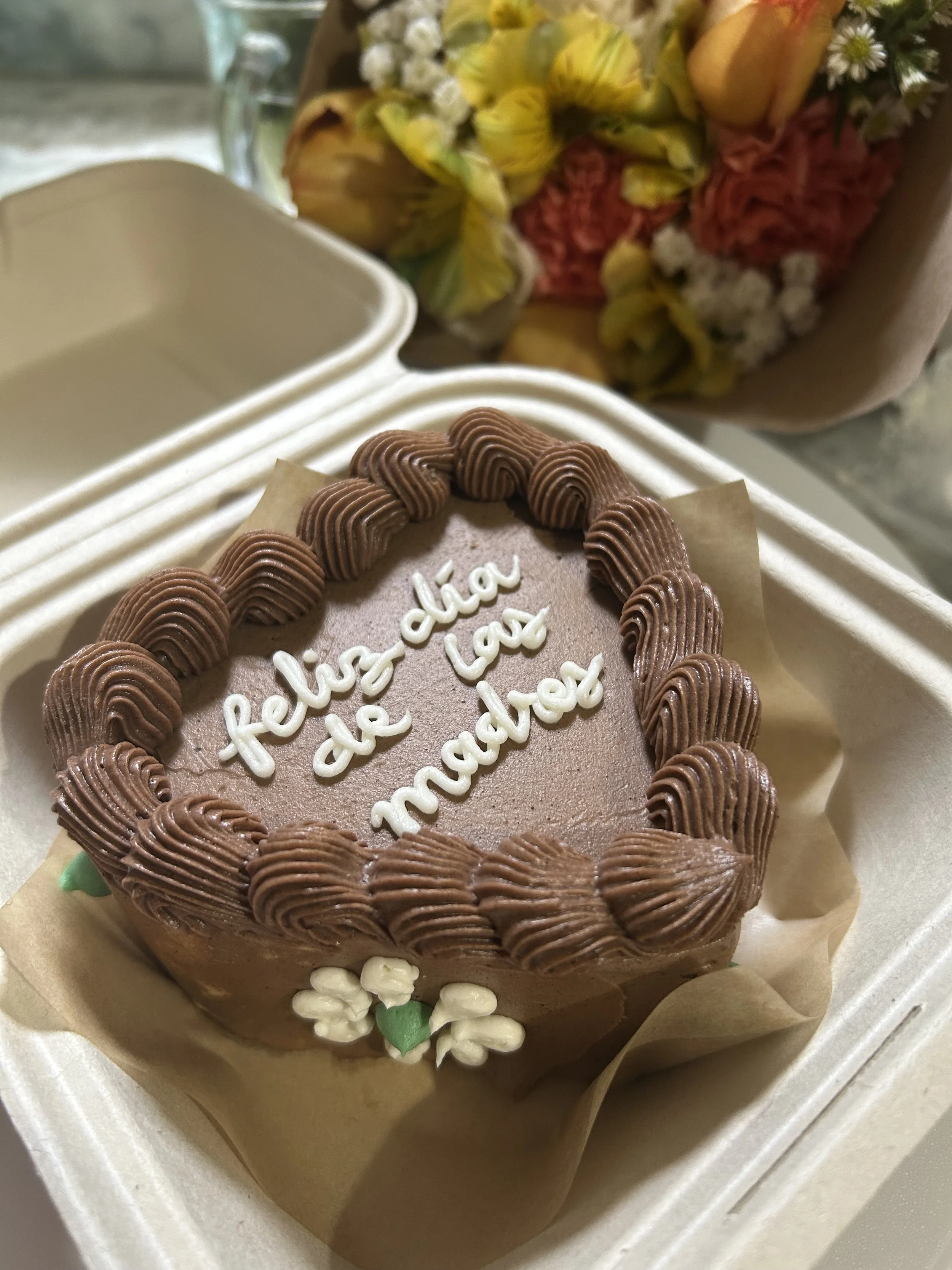 Chocolate cake with a message written in white icing that says "feliz día de madras" inside a cardboard box, with a bouquet of yellow and pink flowers in the background.