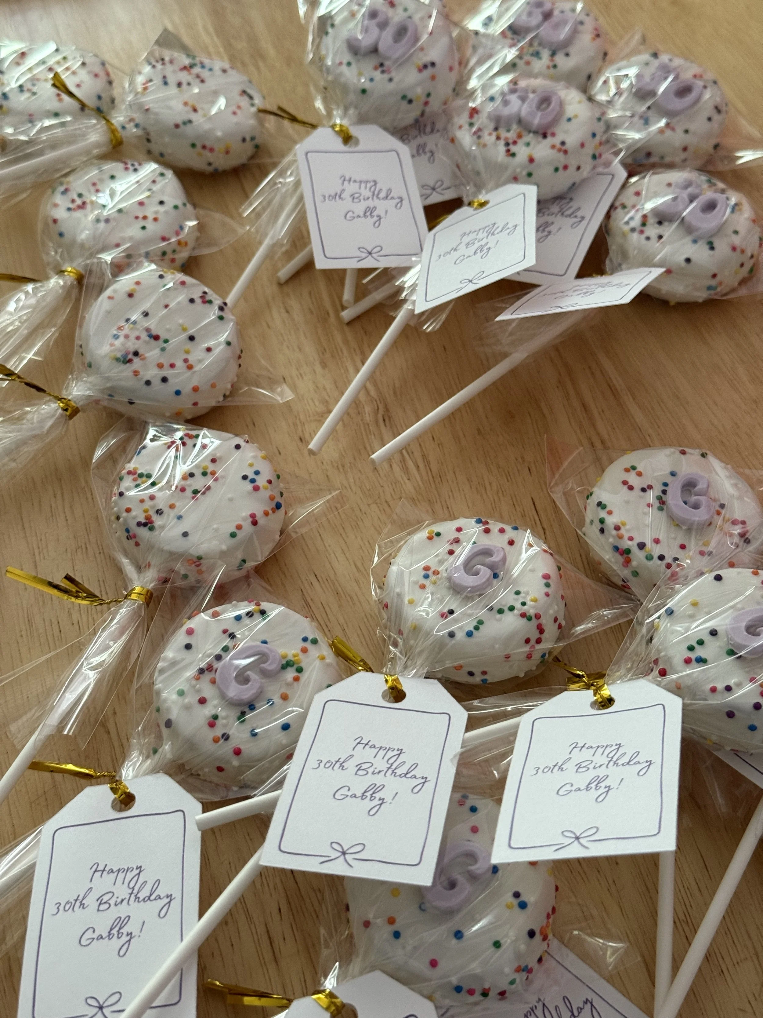 Group of birthday-themed cake pops with white chocolate coating, colorful sprinkles, and a purple number "30" decoration, wrapped in clear plastic and tied with gold ribbons, on a wooden surface.