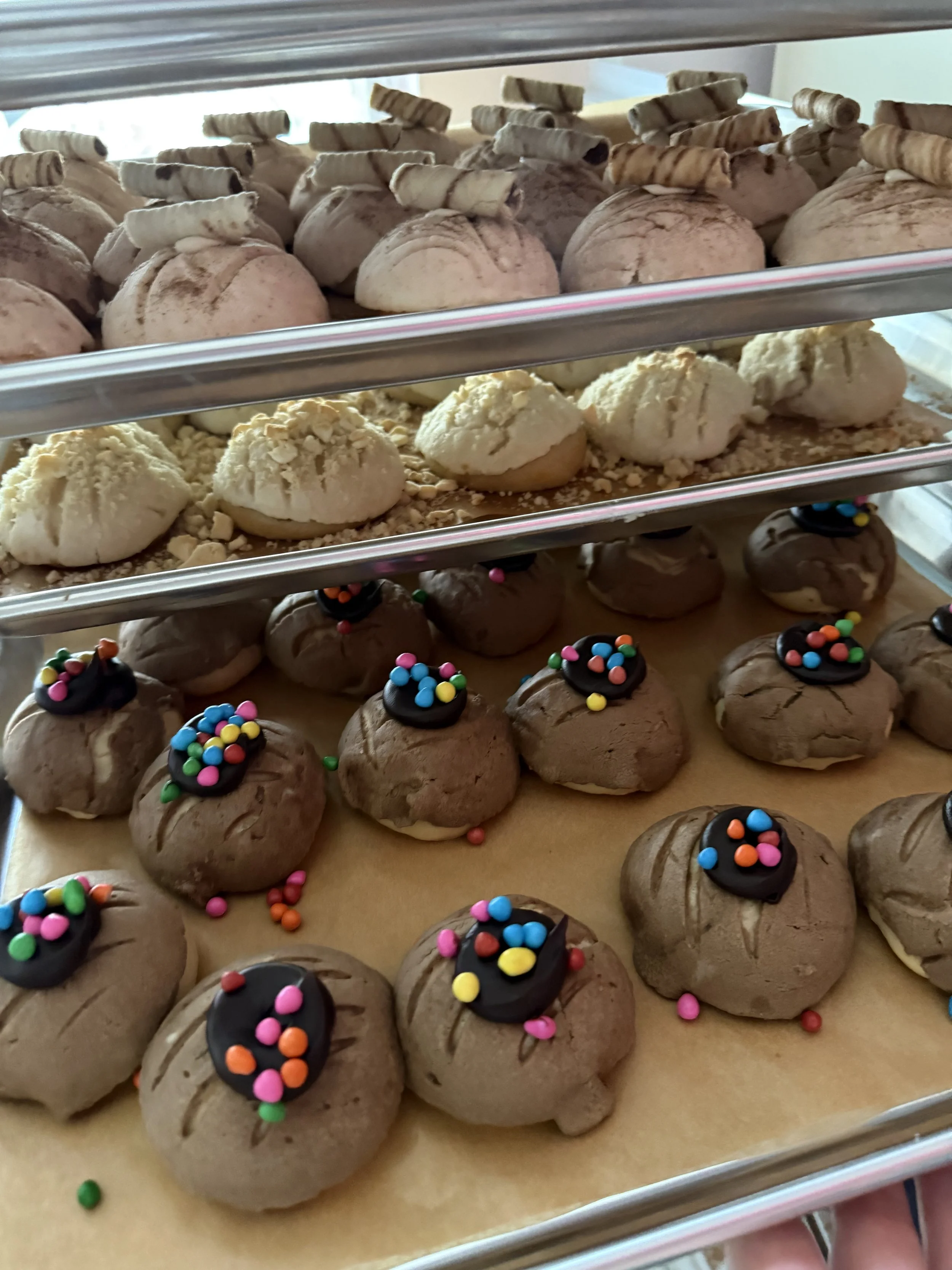 Three shelves of assorted cookies; top shelf has pink cookies with chocolate swirl toppings, middle shelf has pale cookies, bottom shelf has chocolate cookies with colorful sprinkles.