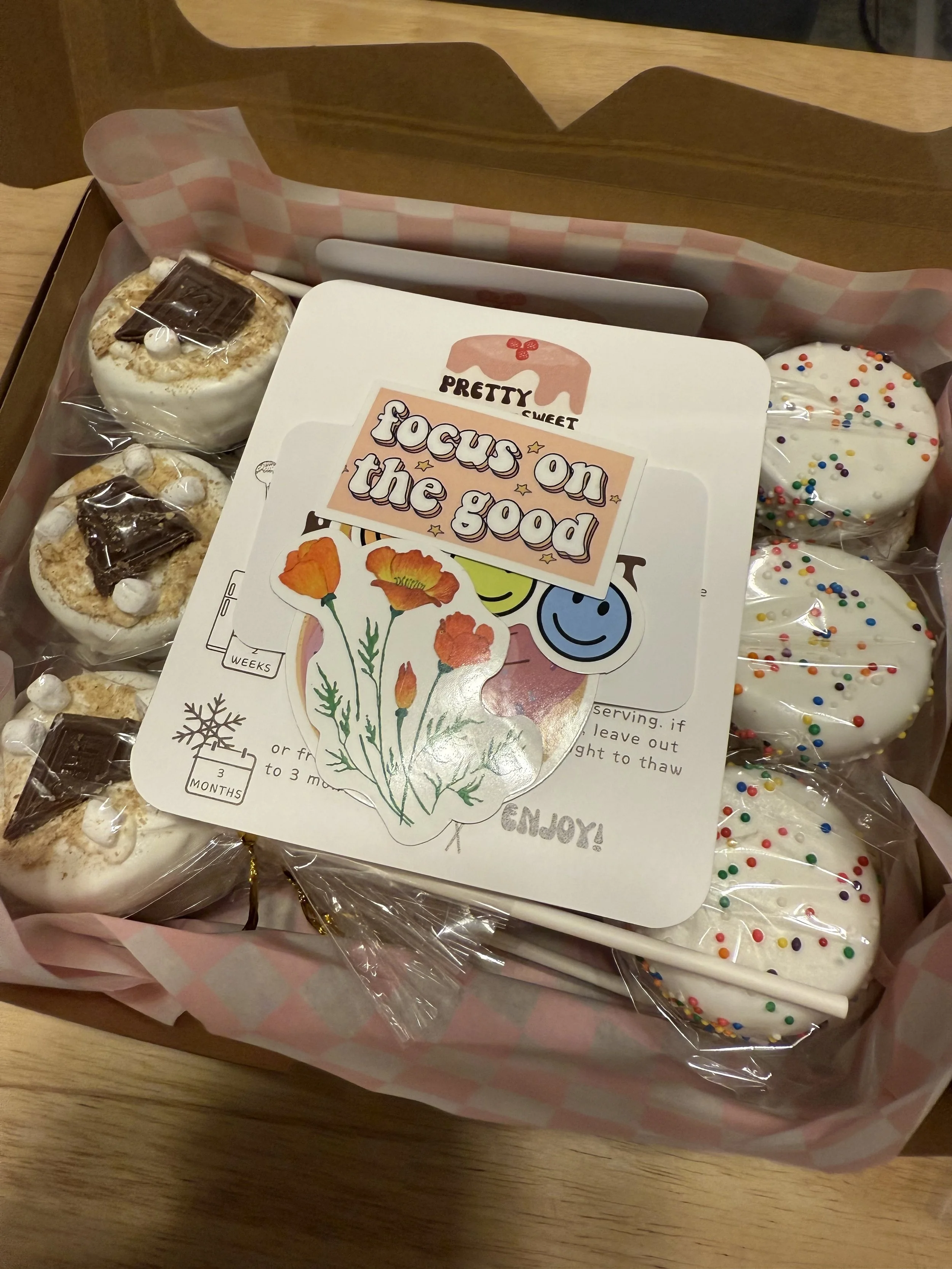 A box of marshmallow treats with colorful sprinkles and chocolate toppings, along with a card that says 'Focus on the good' and decorative stickers of flowers and smiley faces.