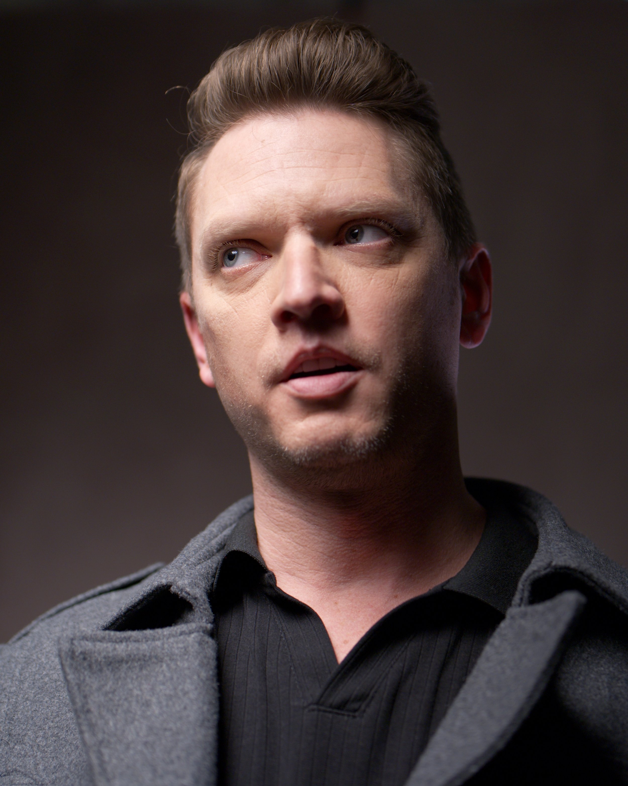 Close-up of a man with light skin, brown hair, and blue eyes, wearing a black shirt and a gray coat, against a dark background.