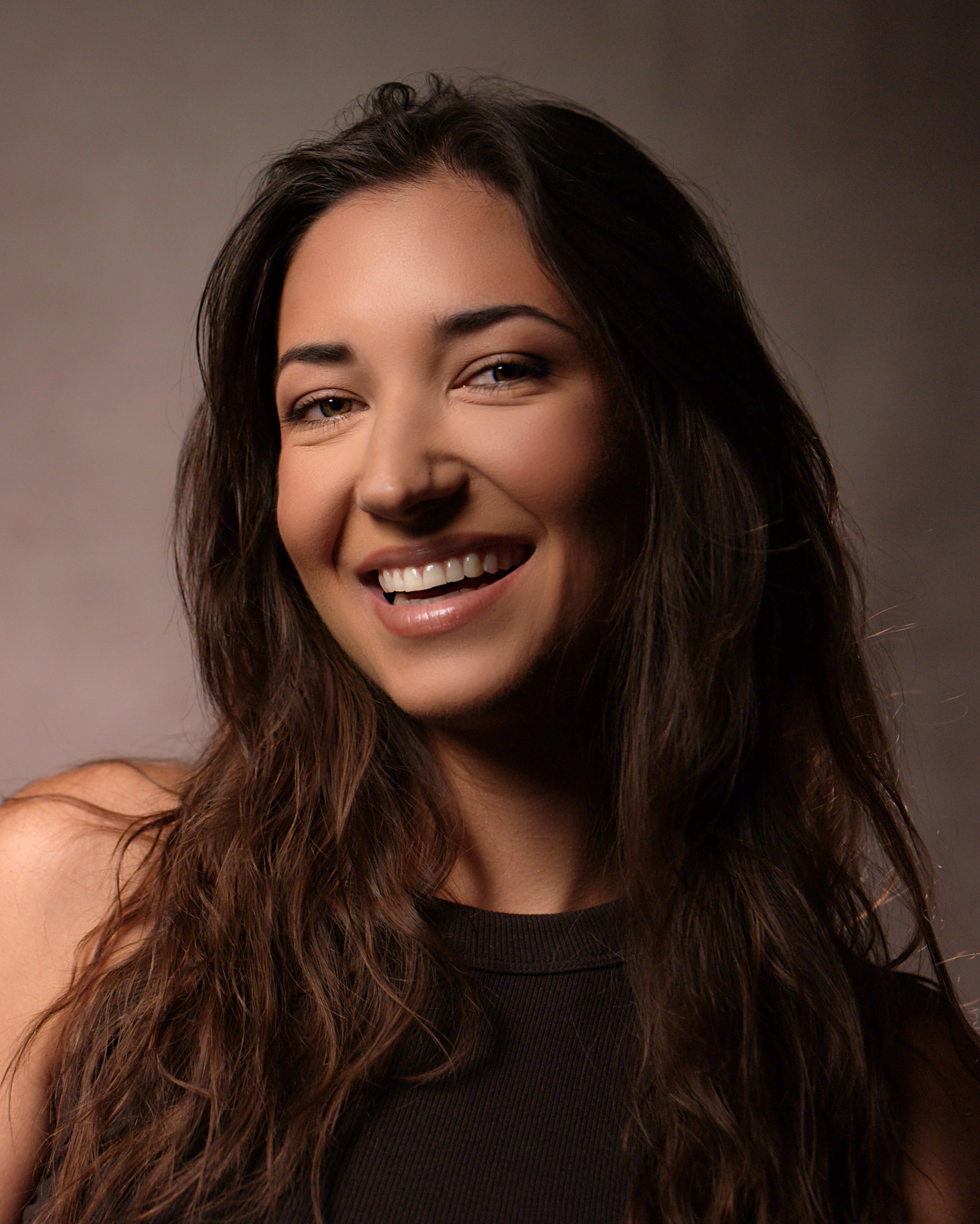 Close-up of a smiling woman with long, wavy dark hair wearing a black sleeveless top against a plain background.