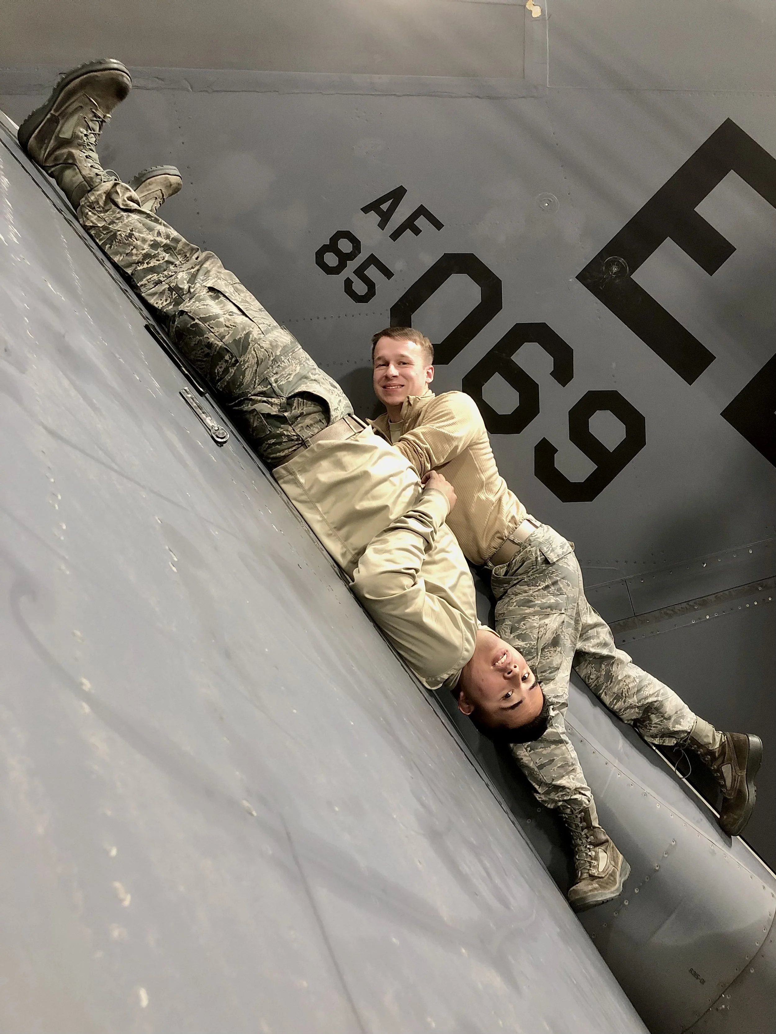 Two soldiers in camouflage uniforms lying on the ground in front of a fighter jet, smiling, with the jet's underbelly and markings visible.