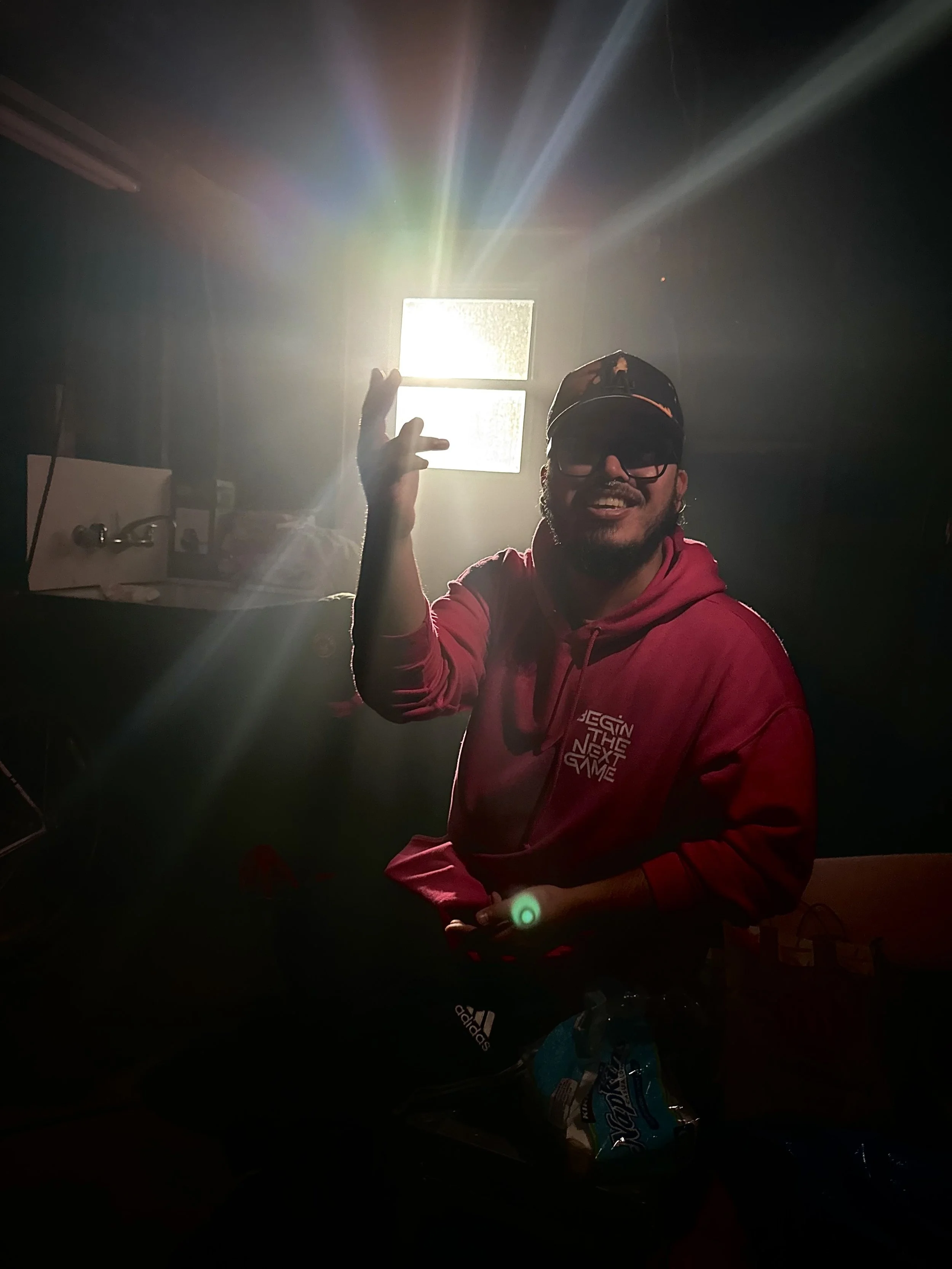 A man in a red hoodie and sunglasses, smiling and making a hand gesture, is illuminated by sunlight coming through a window behind him in a dark room.