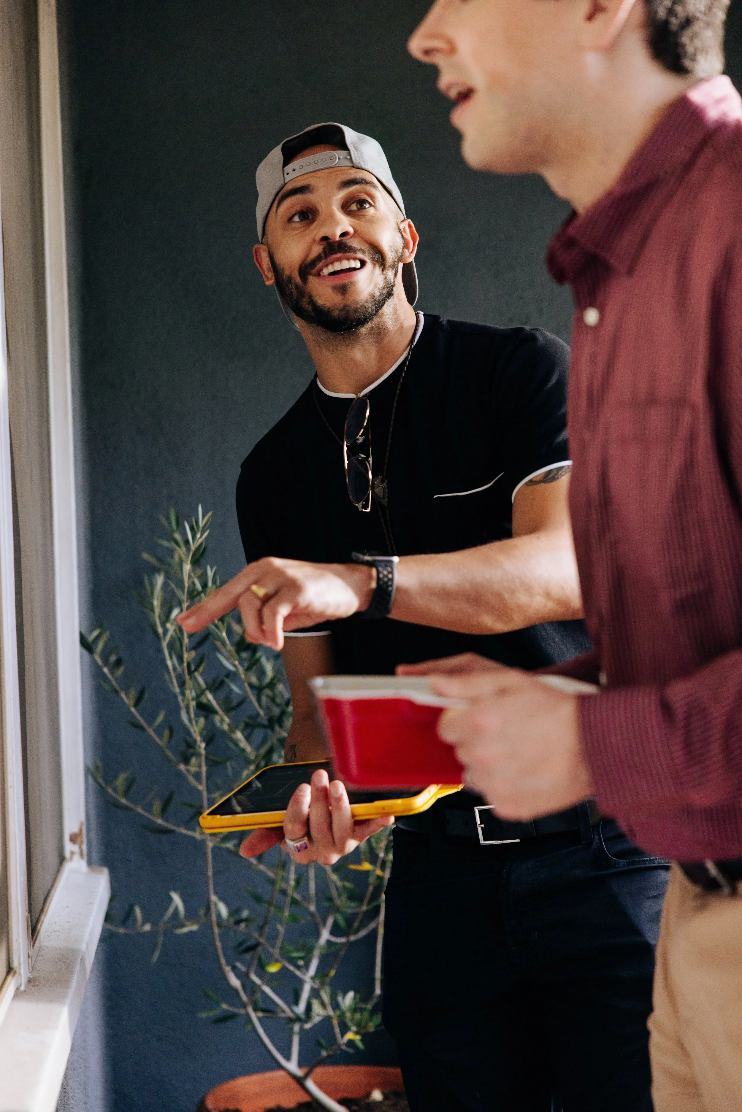 Two men are having a conversation inside a house. One man is holding a smartphone and a red cup, while the other man is pointing and talking animatedly. The man pointing is smiling and wearing a black shirt and a cap.