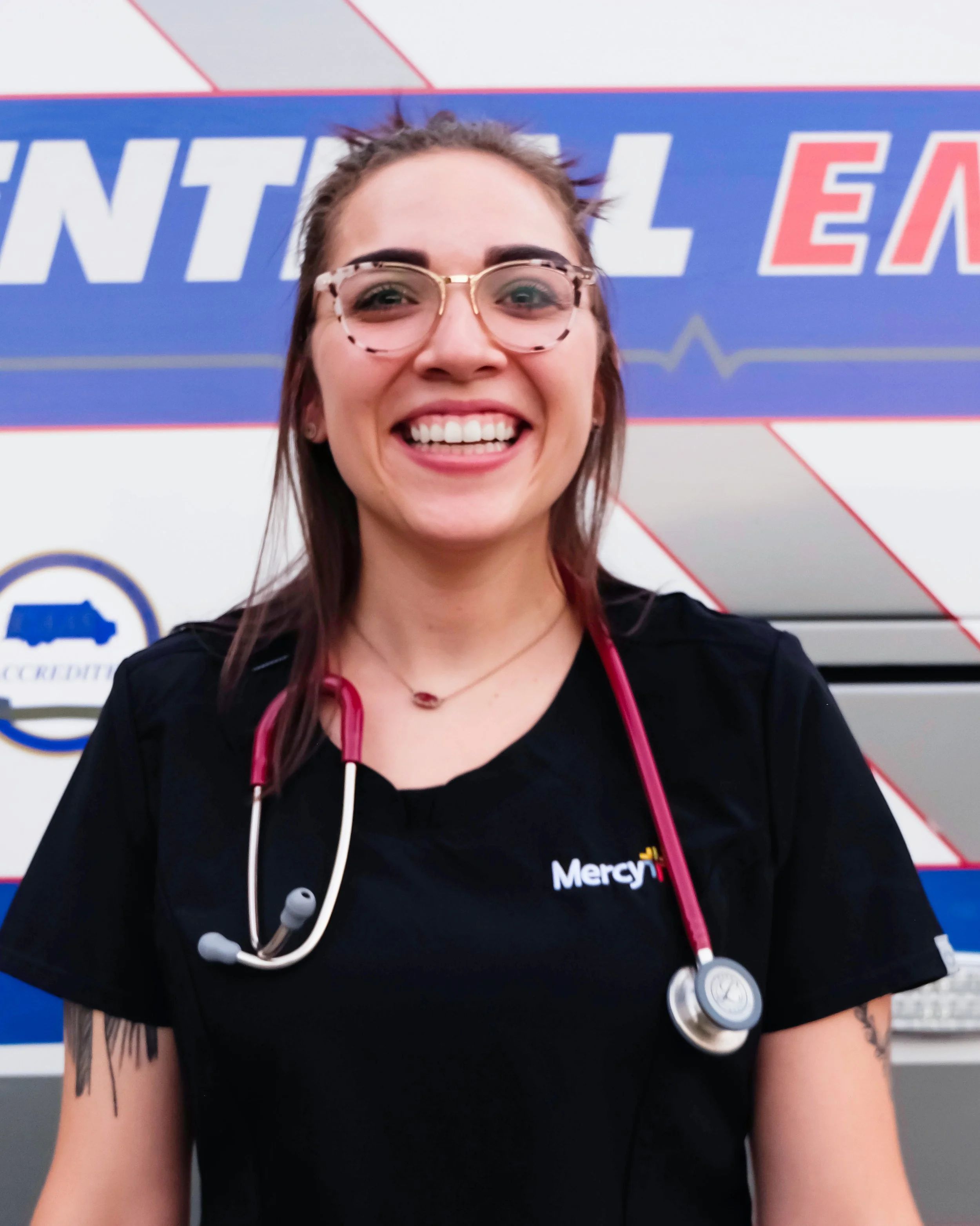 A smiling woman with glasses and a stethoscope around her neck, in front of a healthcare facility sign that reads Mercy.