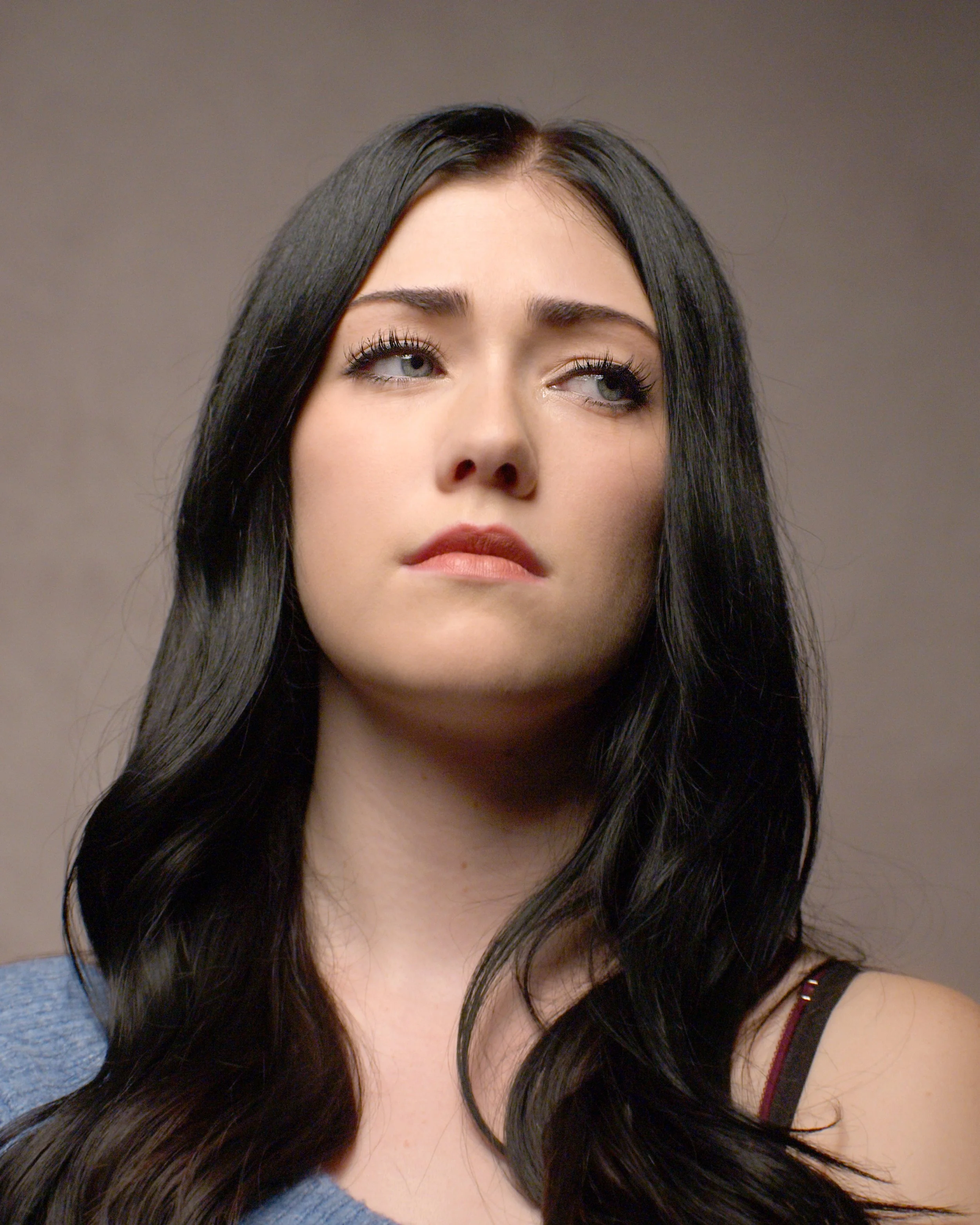 A young woman with long black hair, fair skin, and green eyes is looking slightly upward and to her right. She has makeup on, including mascara and lipstick, and is wearing a black strap top with a thin strap visible on her shoulder.