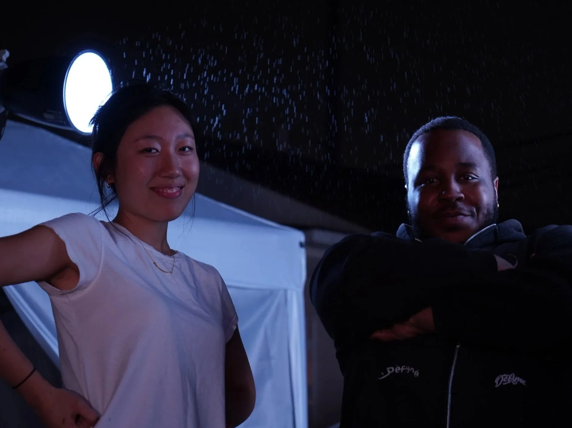 A woman and a man standing under a dark ceiling with water droplets falling around them, illuminated by a bright light in the background.