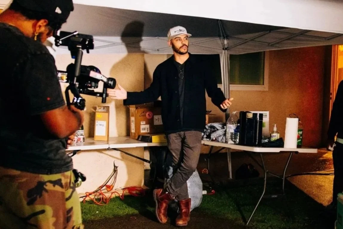 A man in a black jacket, black shirt, and gray pants, wearing a gray cap, stands under a canopy with his arms outstretched. A person with a camera and camouflage pants is filming him. There are boxes, water bottles, and other items on the table behind him.