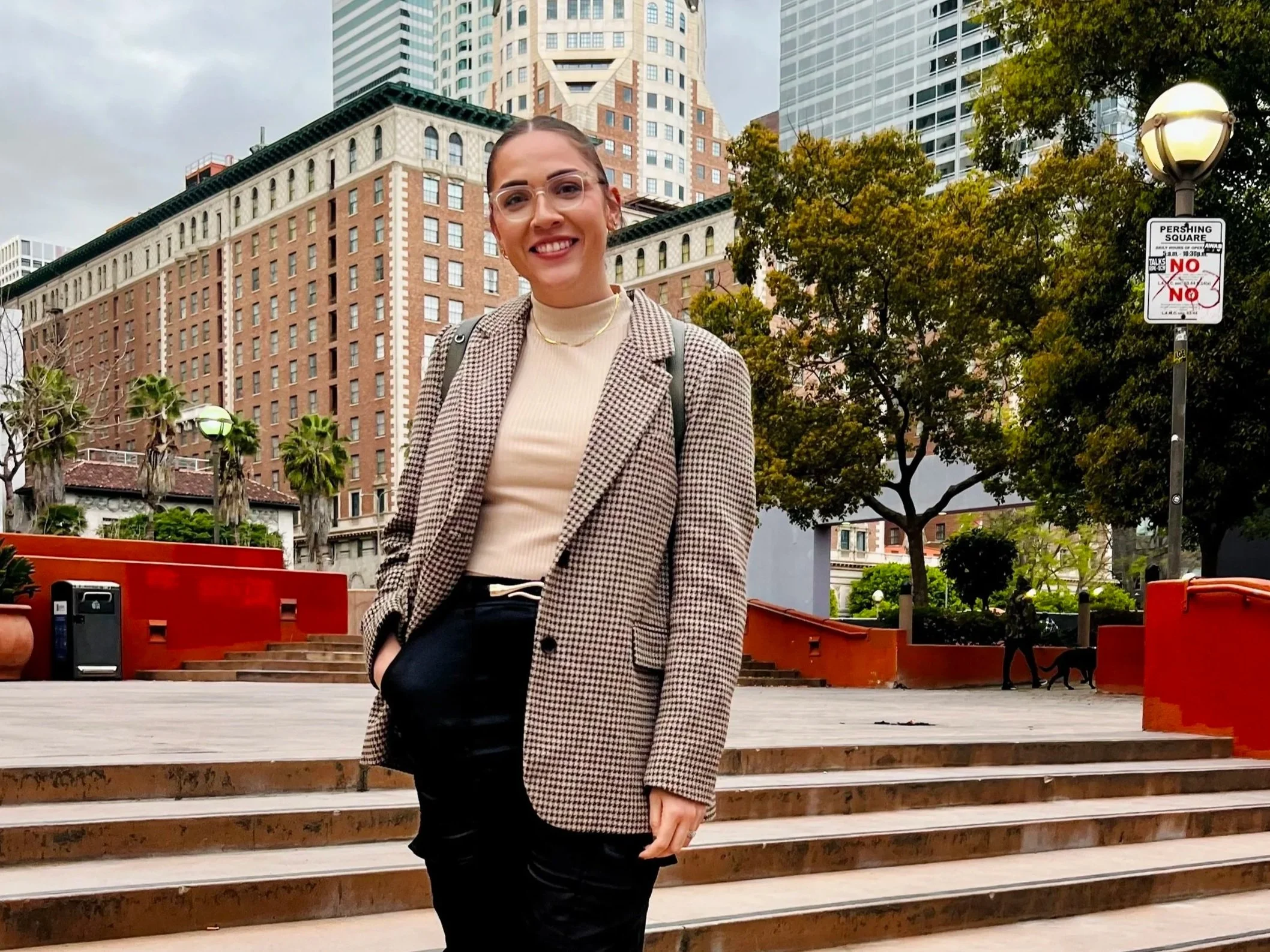 A woman standing behind a metal gate with an 'Emergency Exit Only' sign, wearing a white cap, a black and white plaid shirt, and light gray pants, on a city sidewalk with cars, palm trees, and street signs in the background.
