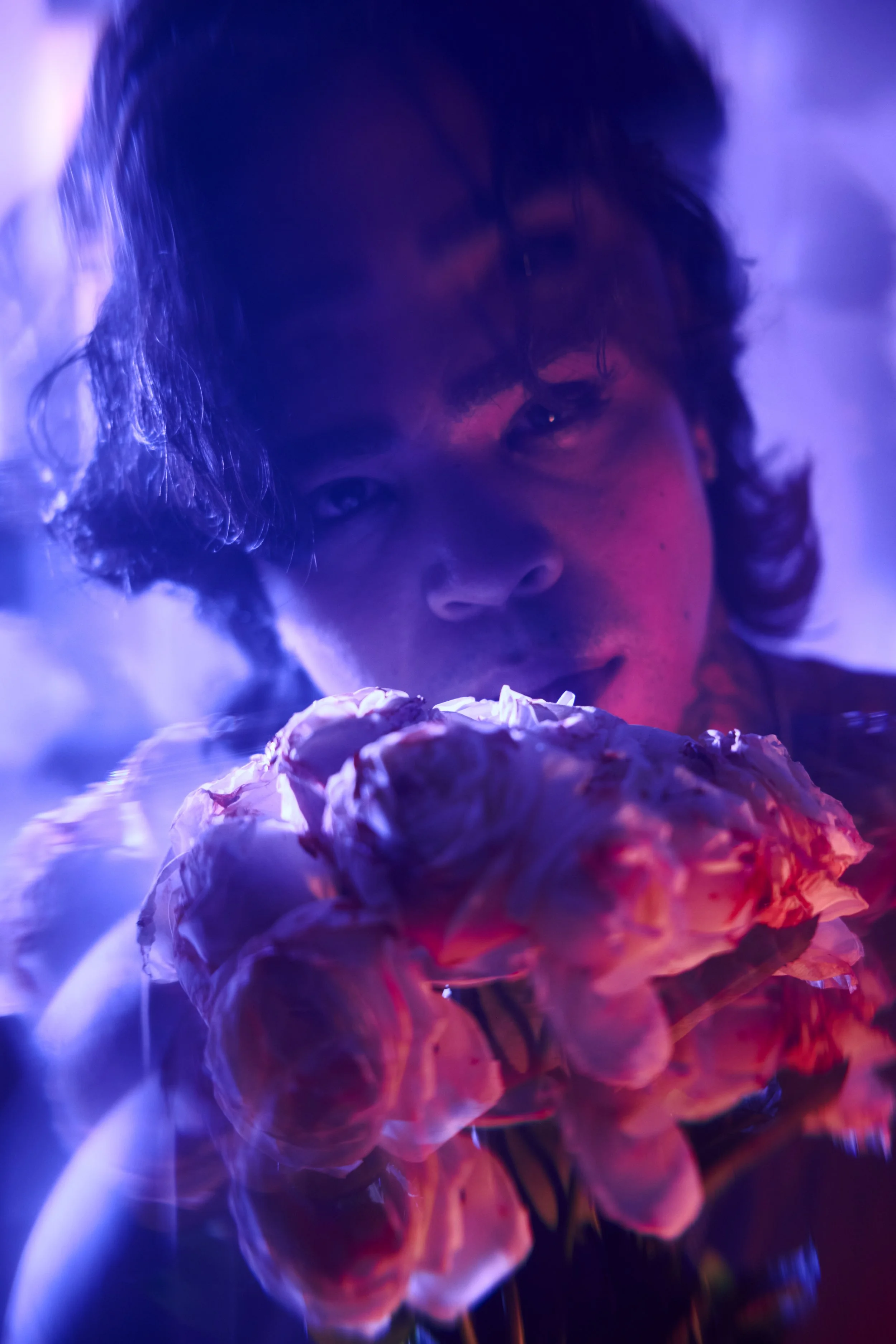 The musical artist, ØUTLR. A person with dark, wavy hair looking at a bunch of pink roses, illuminated by purple and pink lighting.