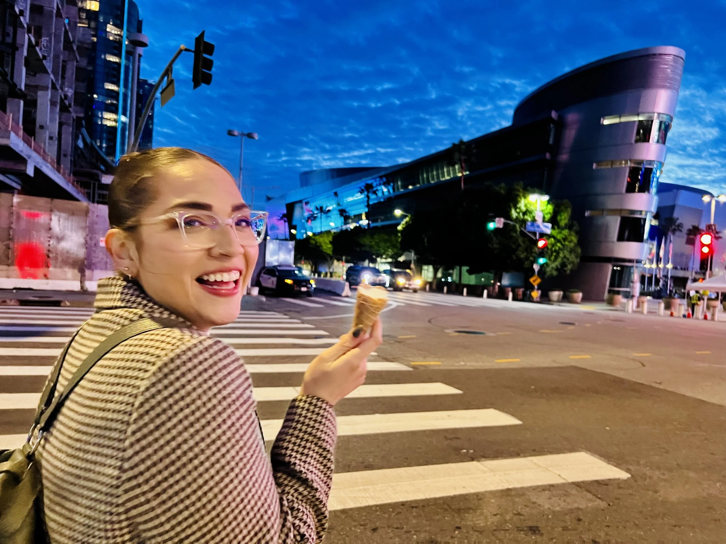 Smiling woman with glasses holding an ice cream cone at a city crosswalk during the evening, with modern buildings and traffic lights in the background.