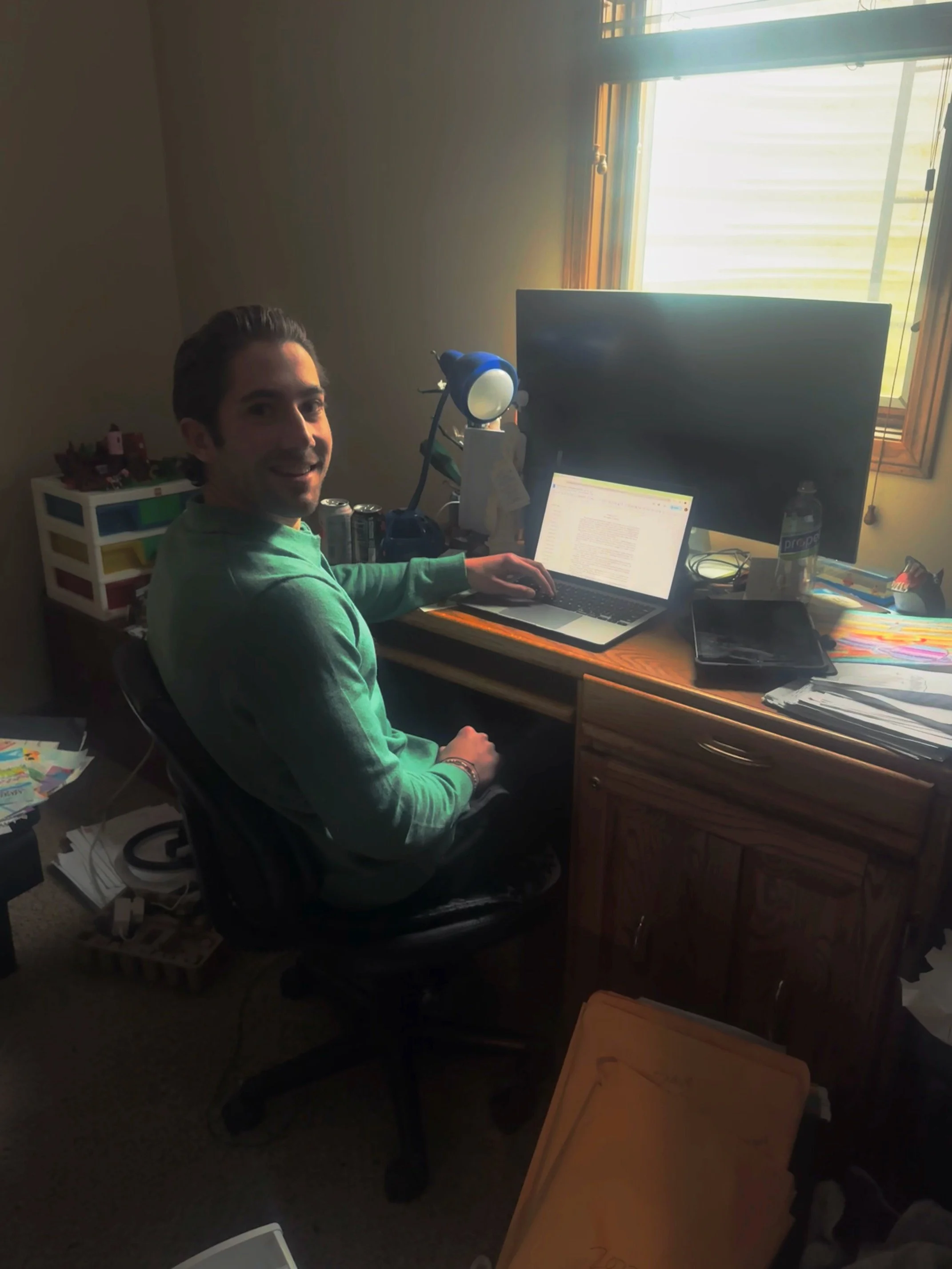 A man sitting at a cluttered wooden desk in a home office, working on a laptop, with a large monitor, a water bottle, and various papers. The room has a window with sunlight coming through.
