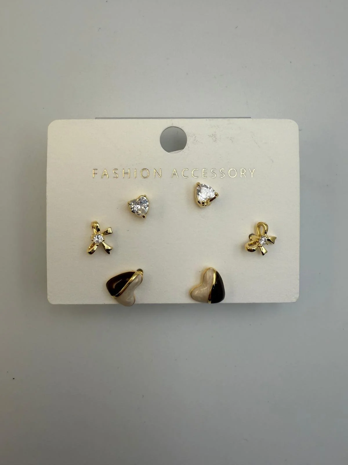 A set of six pairs of fashion earrings on a white display card marked "FASHION ACCESSORY." The earrings include two heart-shaped stud earrings with clear stones, two bow-shaped earrings with small stones, and two abstract-shaped earrings with black, white, and gold accents.