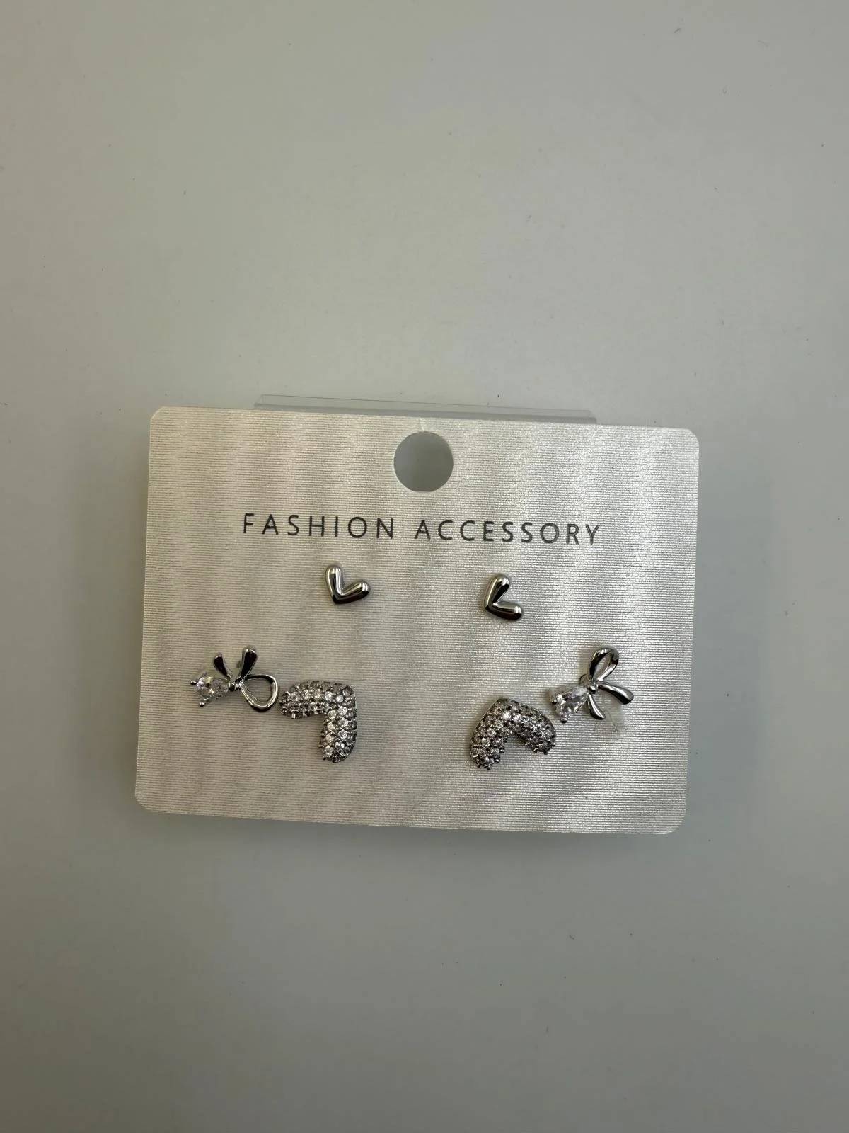 Set of three pairs of earrings on a white card labeled 'Fashion Accessory'. The earrings include two pairs of small silver hoops, and two pairs of decorative earrings with bows and rhinestones.