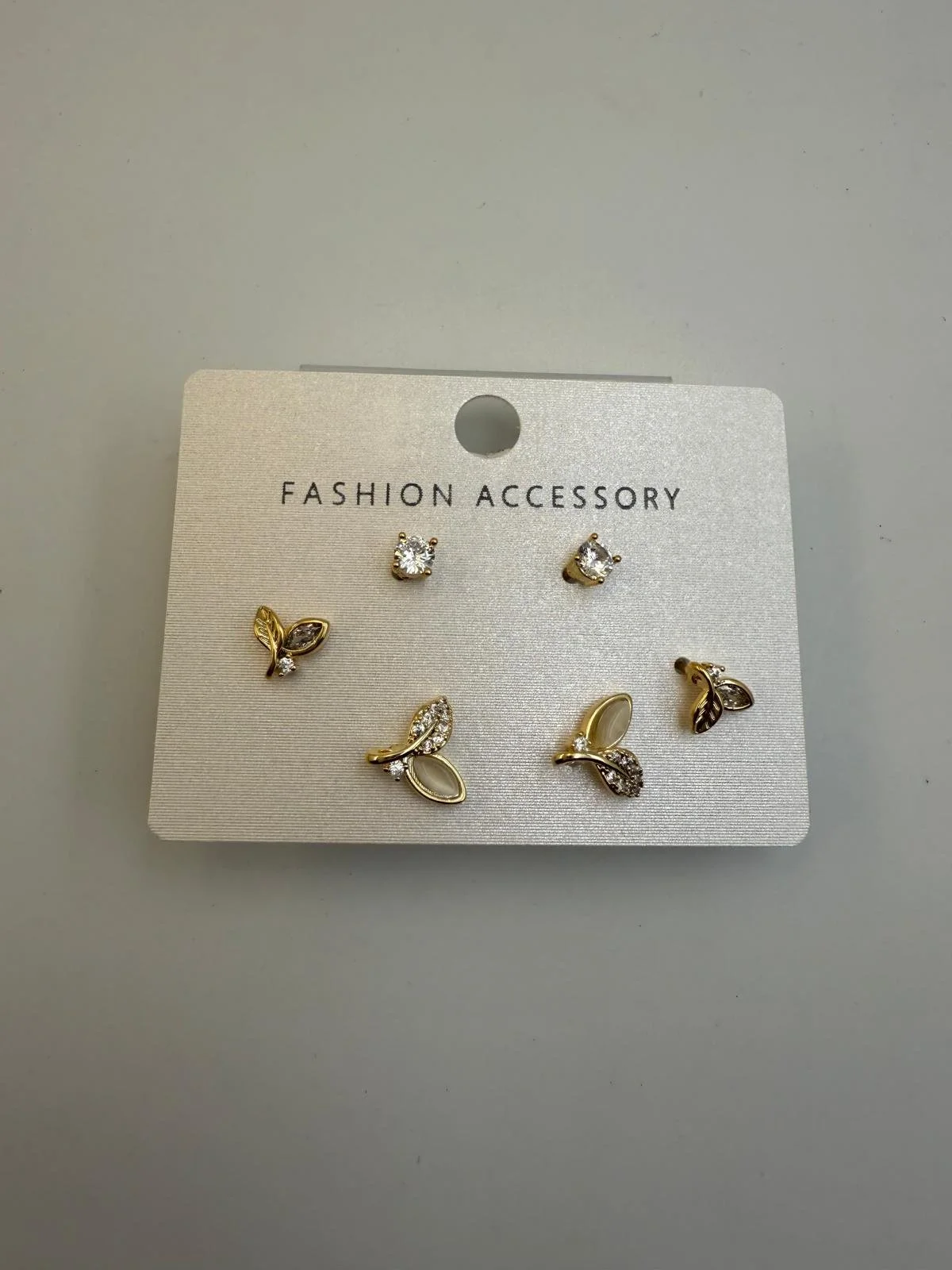 Set of six designer earrings on a gray card labeled "FASHION ACCESSORY". The earrings include pairs of studded round stones and pairs of gold-tone and crystal leaf and flower designs.