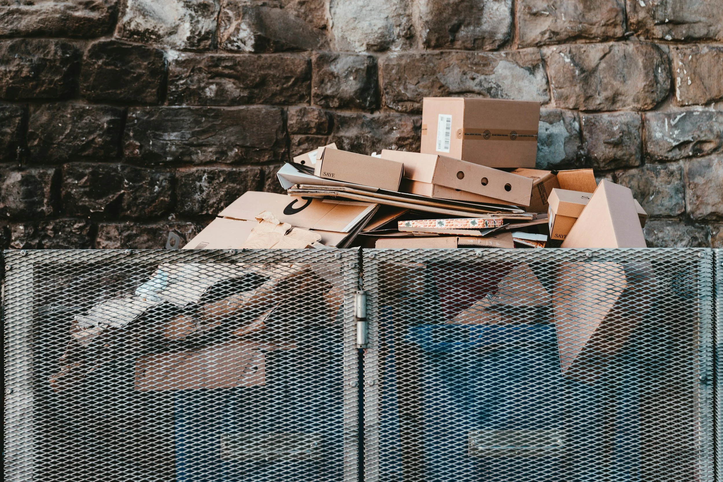 A metal trash or recycling bin filled with flattened cardboard and paper, set in front of a rough stone wall.