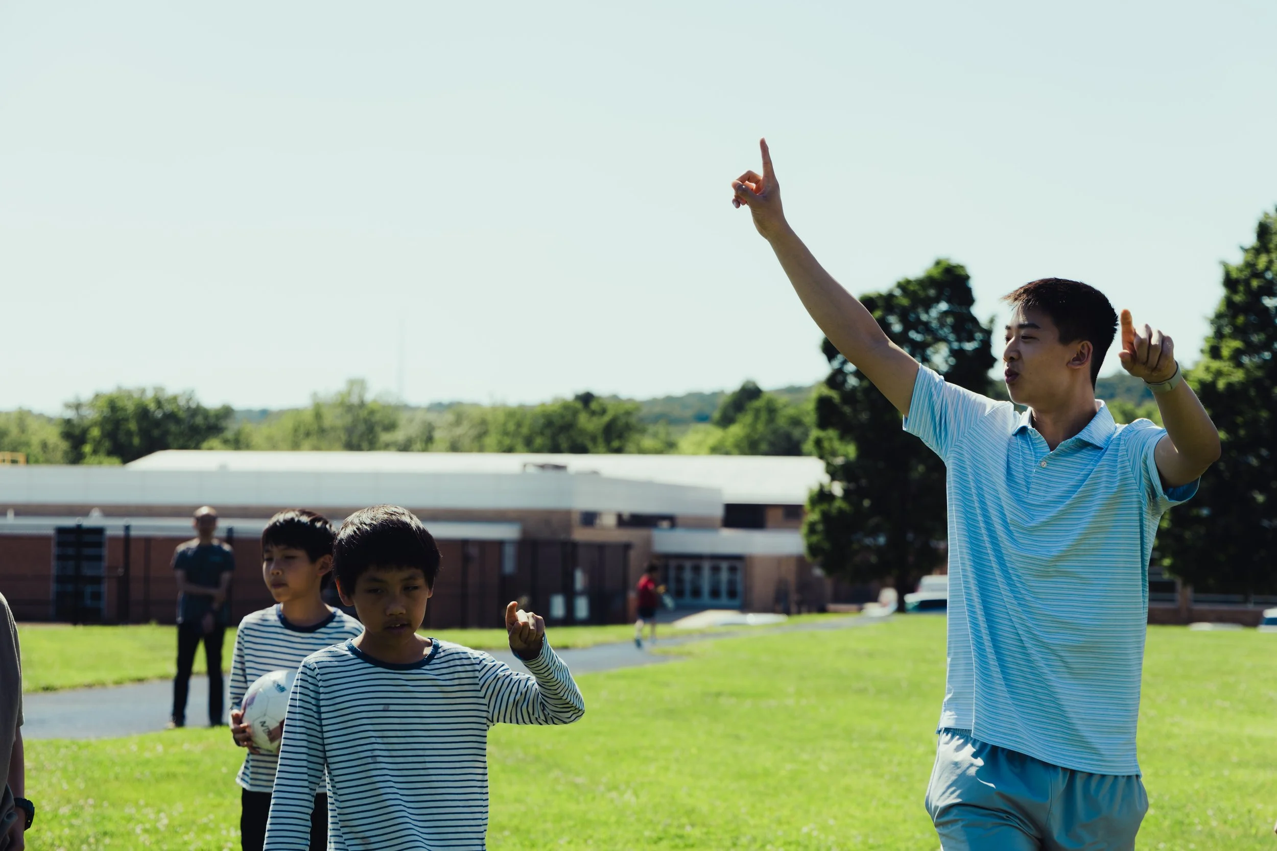 A young man wearing a blue striped shirt gestures upward with his index finger while standing outdoors on a grassy field with children around him during daytime.