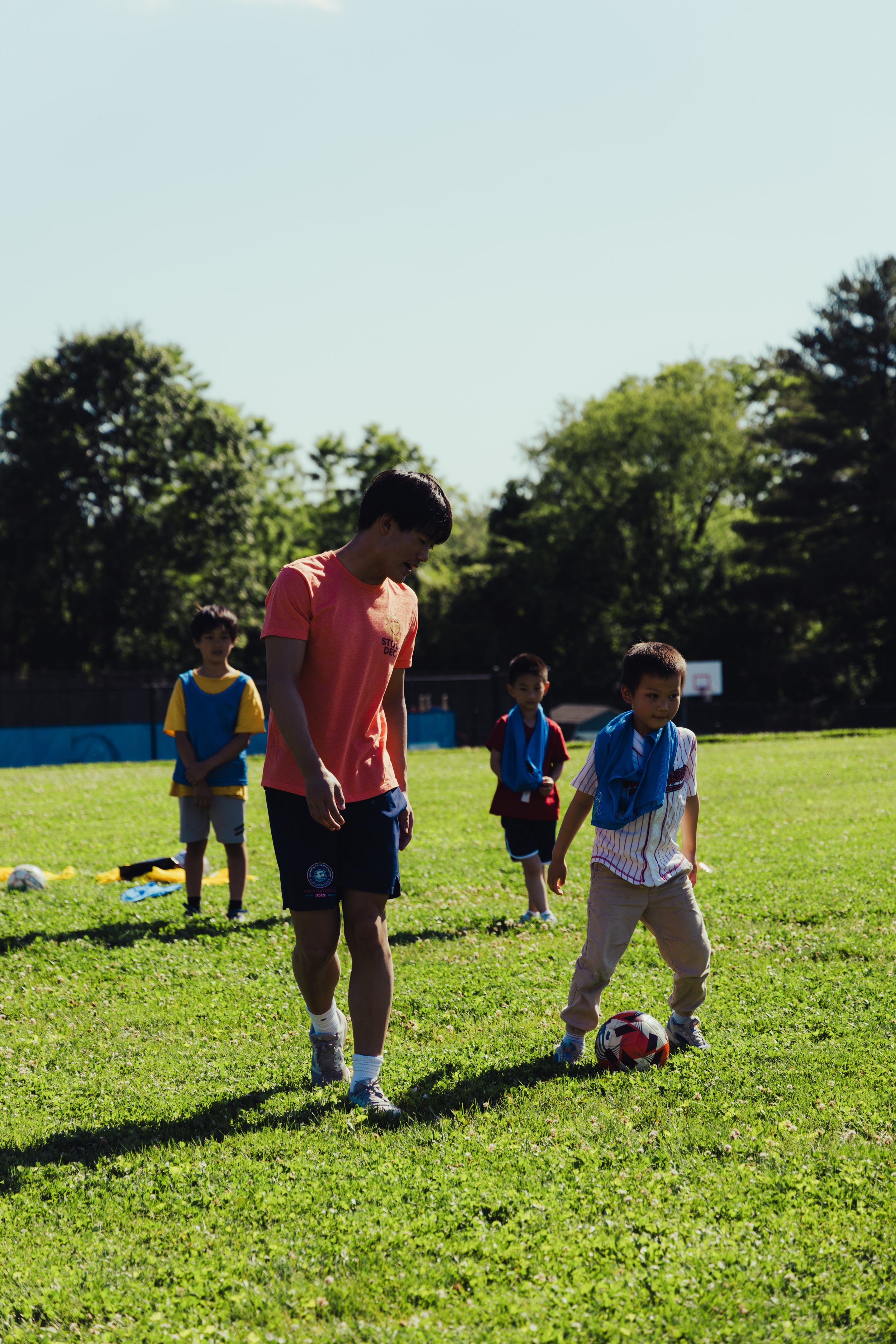 Children playing soccer on a grassy field on a sunny day, with trees in the background.