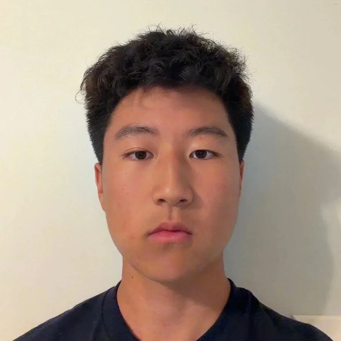 A young man with short, curly black hair, light skin, and wearing a black shirt, standing against a plain white wall.