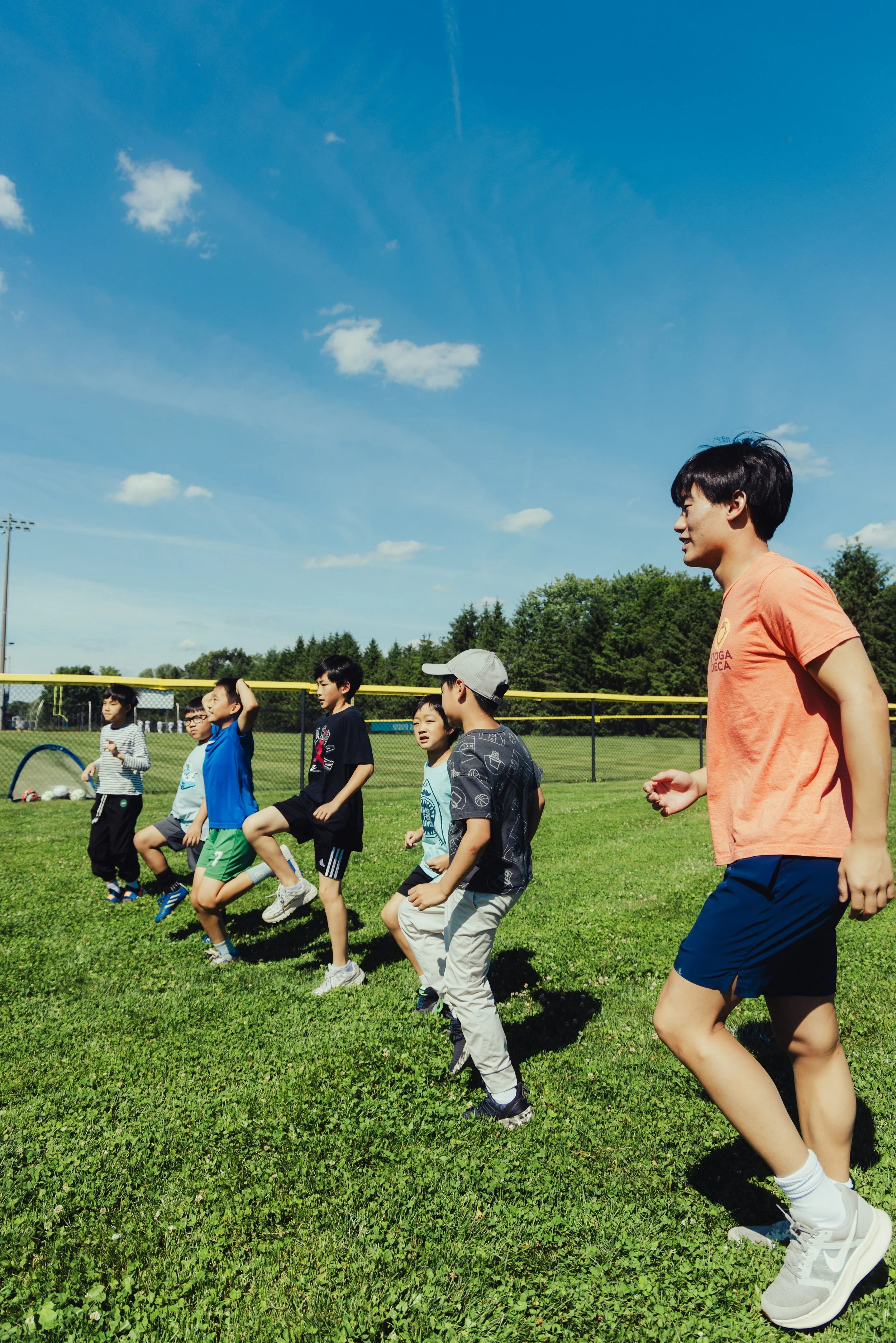 A group of children and an adult participating in outdoor exercise or a fitness activity on a grassy field with a blue sky and some clouds overhead.