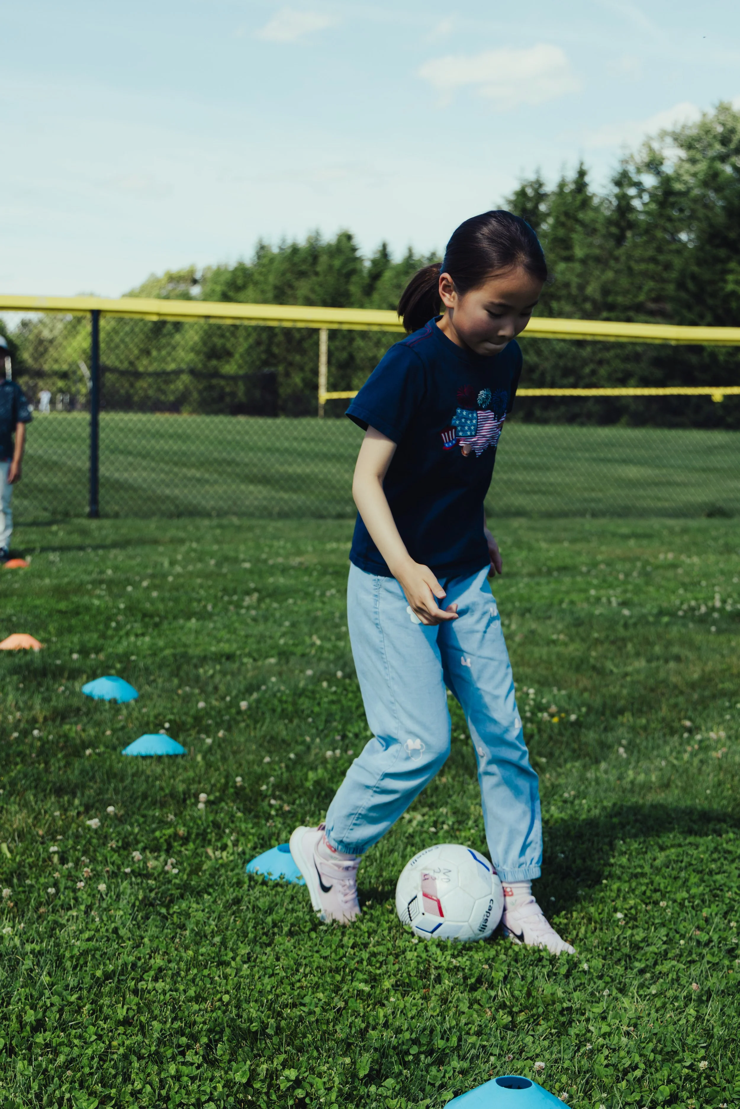 A young girl playing soccer outdoors on a grassy field with cones and a soccer ball, with a yellow fence and trees in the background under a partly cloudy sky.