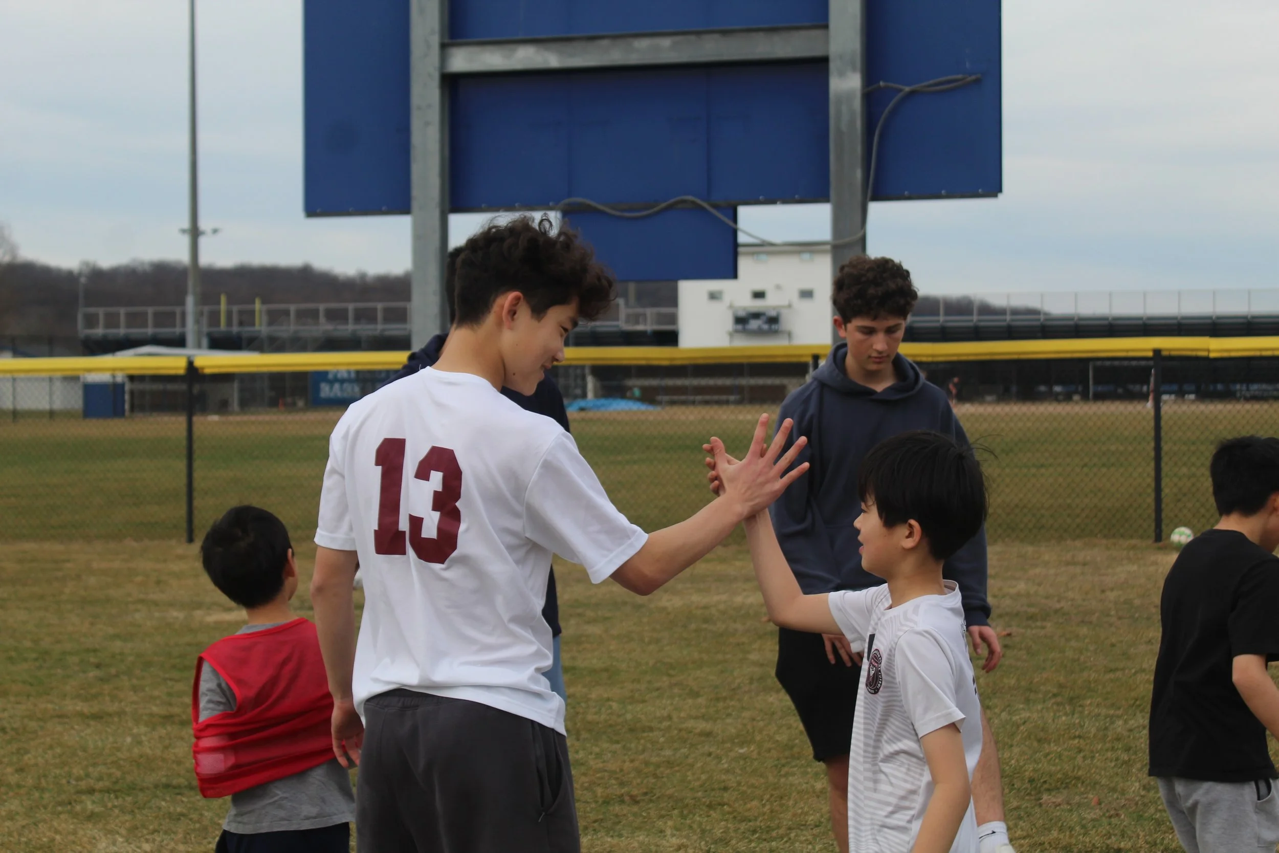 A group of young boys gathered on a football field, with one wearing a white jersey numbered 13, giving a high-five to a kid in a white shirt. There are two older individuals in the background, and a large blue scoreboard behind them. The scene appears to be an outdoor sports practice or game, with a cloudy sky overhead.